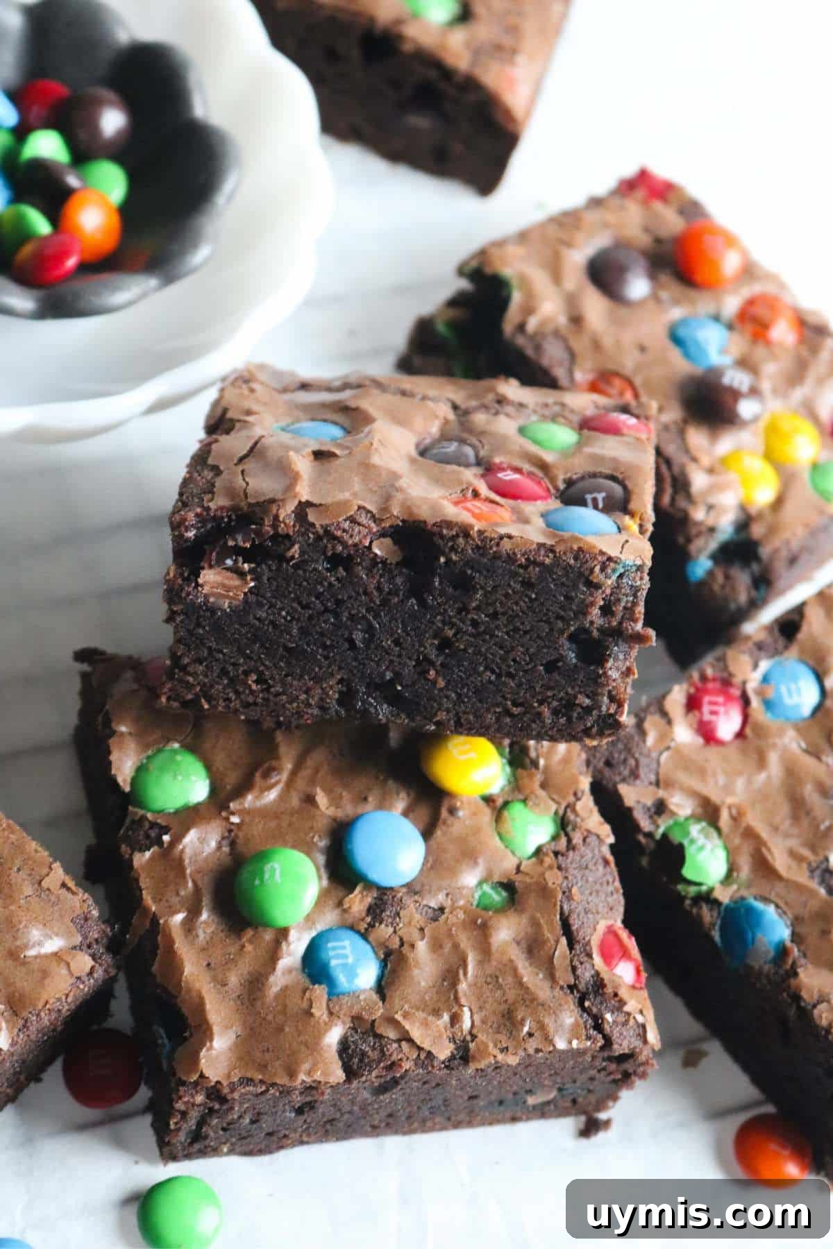 Thick and Chewy M&M Brownies Recipe 11 stack of homemade m&ms brownies shingled together on a white surface with a bowl of candies in the background.