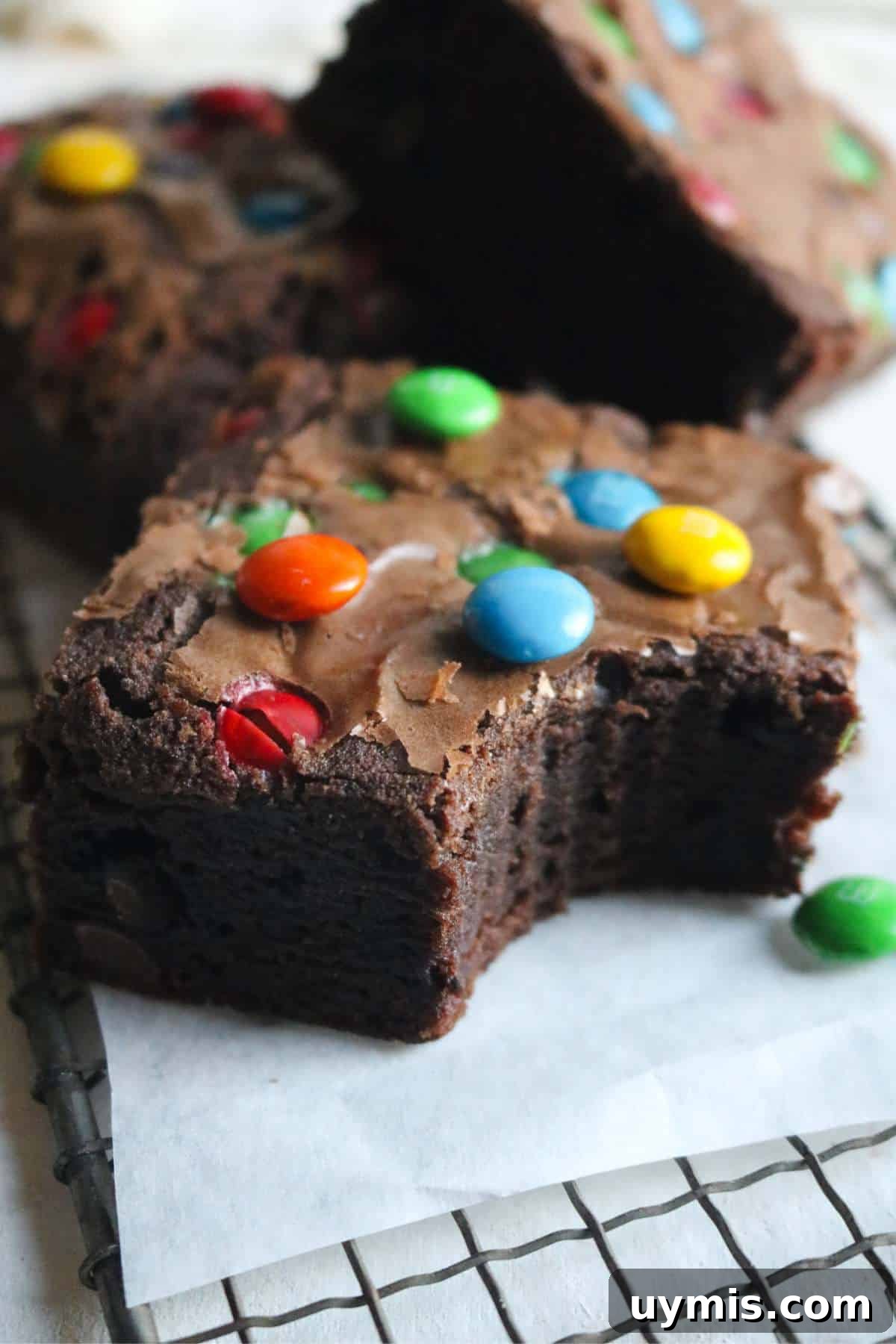 Thick and Chewy M&M Brownies Recipe 3 close up view of a thick brownie studded with m&ms with more brownies shingled in the background.