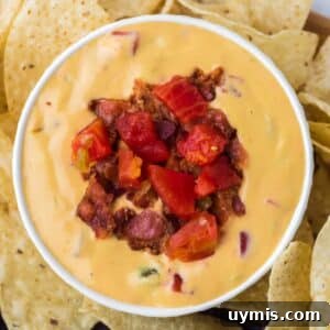 A white bowl filled with nacho cheese dip that's been garnished with bacon bits and tomato. Served with tortilla chips around the outside.