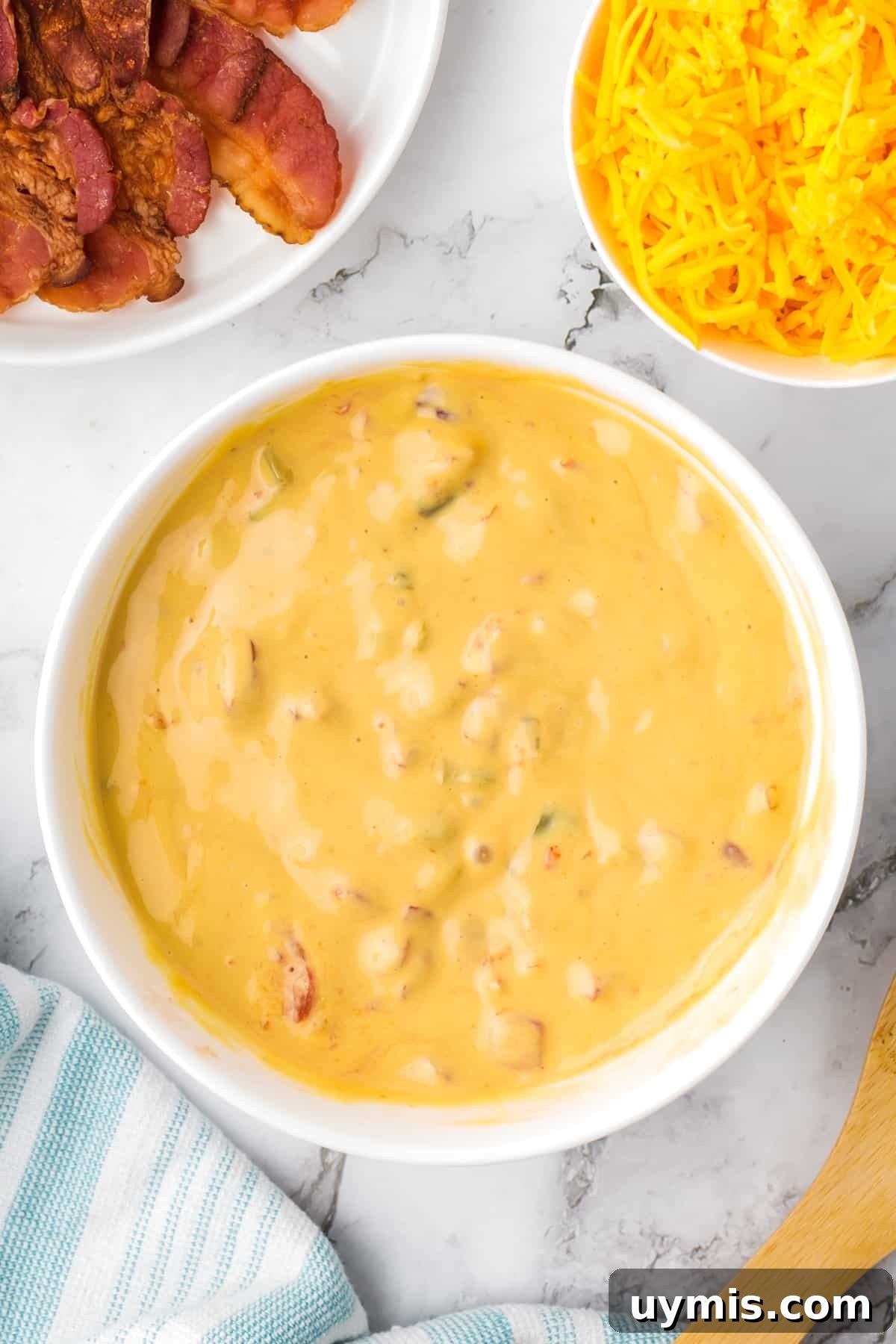 Homemade Nacho Cheese Dip Recipe 6 A large bowl of melted nacho cheese dip, with bowls of shredded cheese and bacon on the side.
