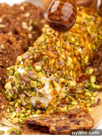 A honey stick drizzling honey on a goat cheese log covered in pistachios.