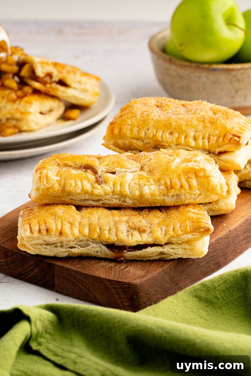 Warm Apple Hand Pies with Cinnamon and Flaky Crust 7 A bunch of apple hand pies stacked on top of a wood cutting board with a bowl of granny smith apples on the table behind them.