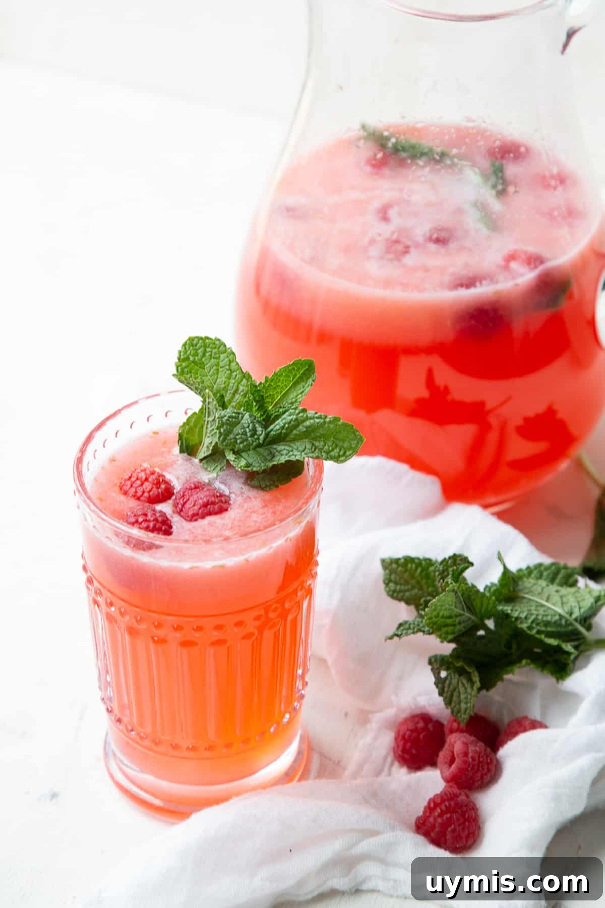 Two Ingredient Raspberry Lemonade Recipe 2 glass of raspberry lemonade garnished with mint, next to a pitcher of more raspberry lemonade.