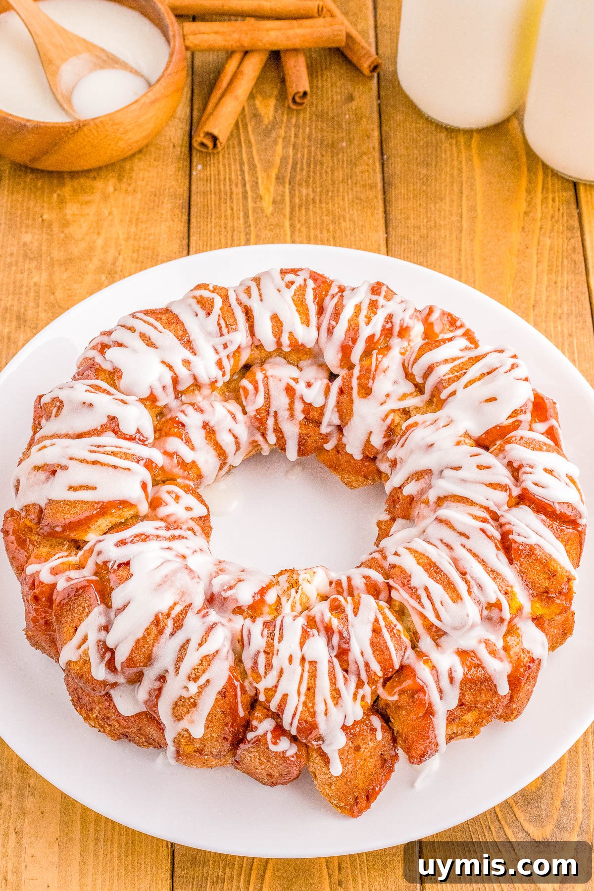 Pull-Apart Cinnamon Roll Monkey Bread Recipe 3 Cinnamon roll monkey bread with white icing drizzle on a white plate, surrounded by cinnamon sticks, a bowl of sugar, and a glass of milk on a wooden table.