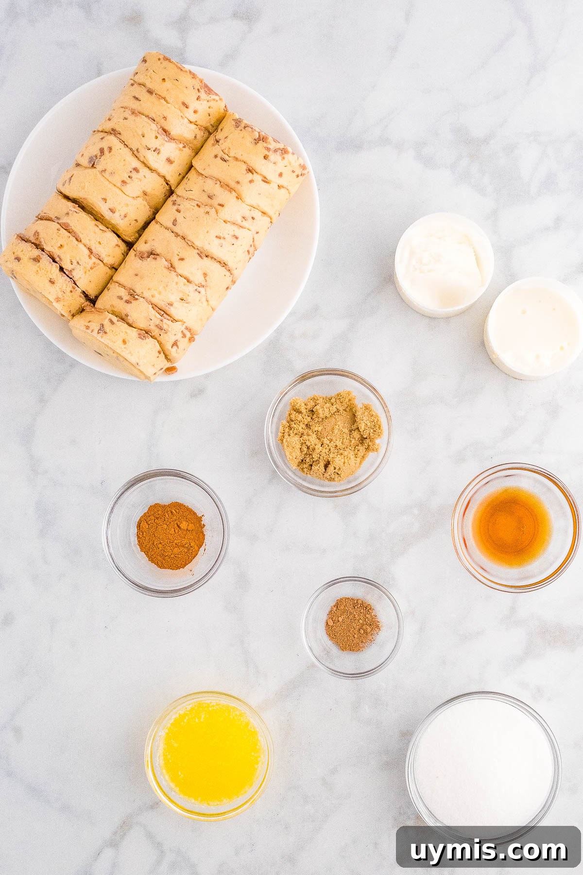 Pull-Apart Cinnamon Roll Monkey Bread Recipe 5 Ingredients for baking on a marble surface, including a plate of cookies, bowls of flour, spices, and sugar, and small containers of honey and vanilla extract.