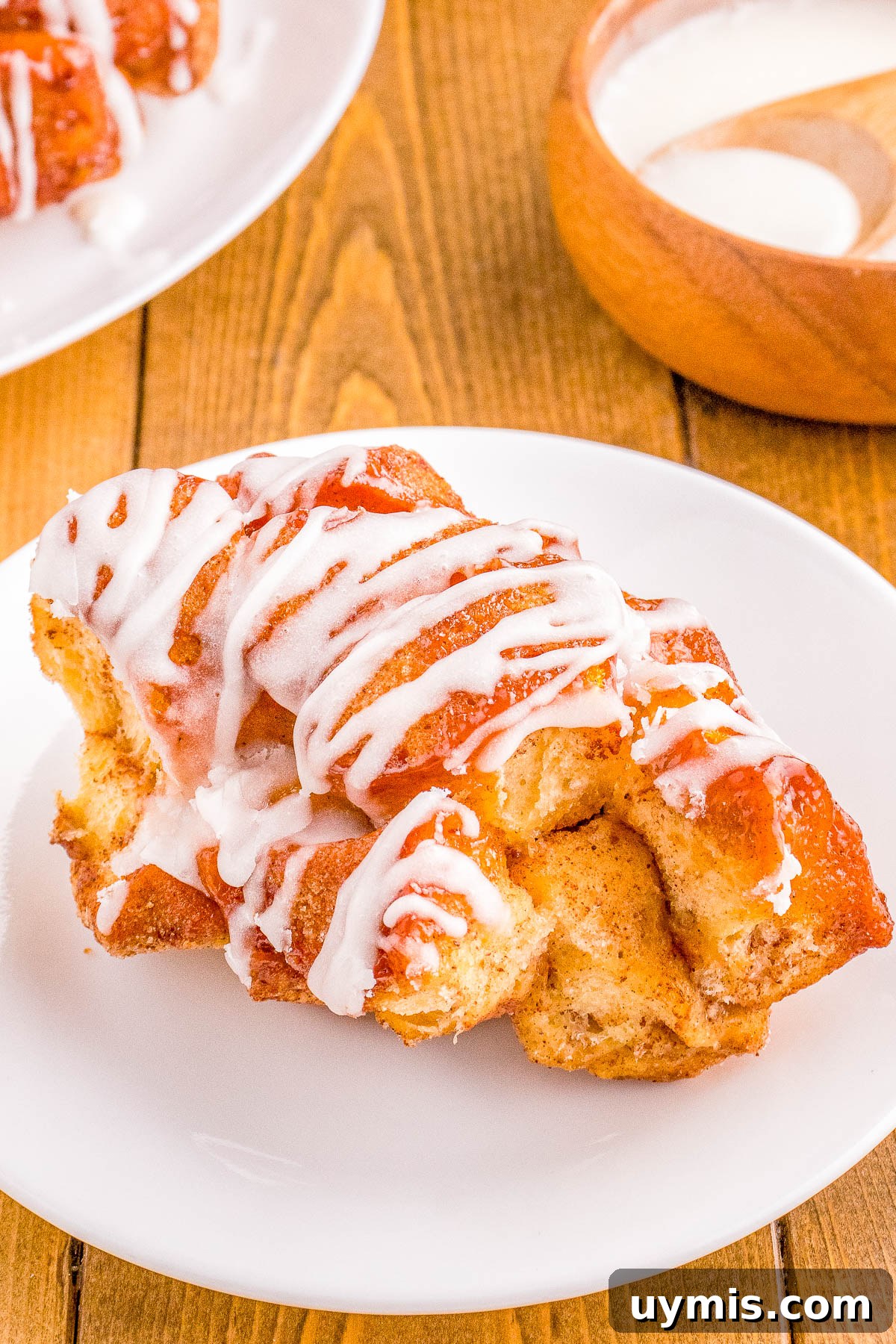 Pull-Apart Cinnamon Roll Monkey Bread Recipe 6 A plate containing a freshly baked cinnamon roll drizzled with white icing, on a wooden table with a bowl of extra icing in the background.