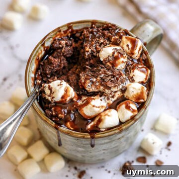 hot chocolate mug cake, covered in marshmallows and chocolate syrup, with a spoon.