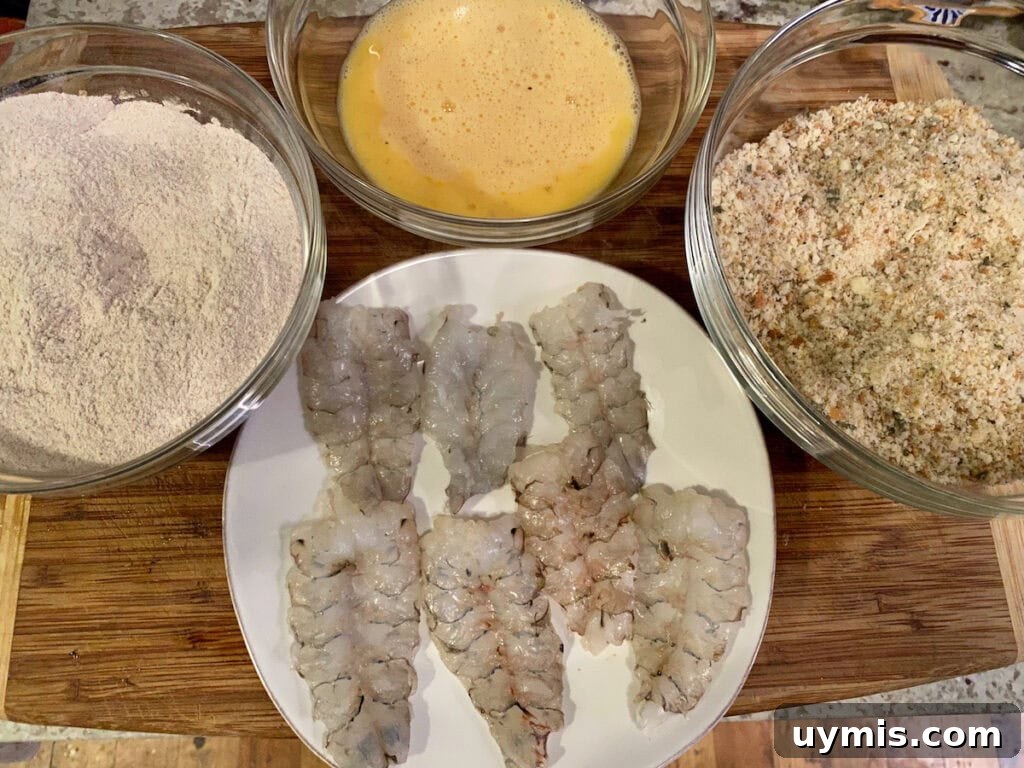 Italian Pan Fried Butterflied Shrimp Recipe 4 Butterflied shrimp on white plate next to bowls of flour, beaten eggs and breadcrumb mixture.