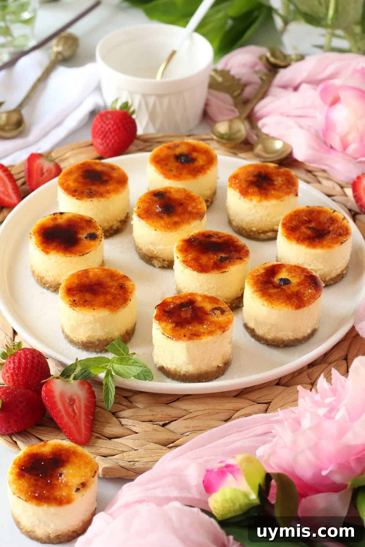 Mini Crème Brûlée Cheesecake Bites Recipe 5 Many creme brulee mini cheesecake with caramelized tops on white plate with strawberries and mint leaves next to it.