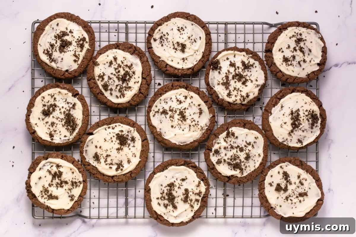 27 Chocolate Desserts You Can't Keep at Home 15 A cooling rack with twelve Crumbl Copycat Frosted Oreo Cookies topped with white frosting and chocolate crumbs, arranged on a white marble surface.