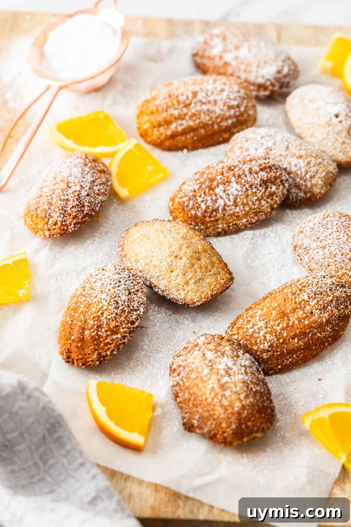 Spiced Orange Madeleines Recipe 2 Spiced Orange Madeleines on some baking paper on a wooden board, dusted with icing sugar and a few pieces of fresh orange.