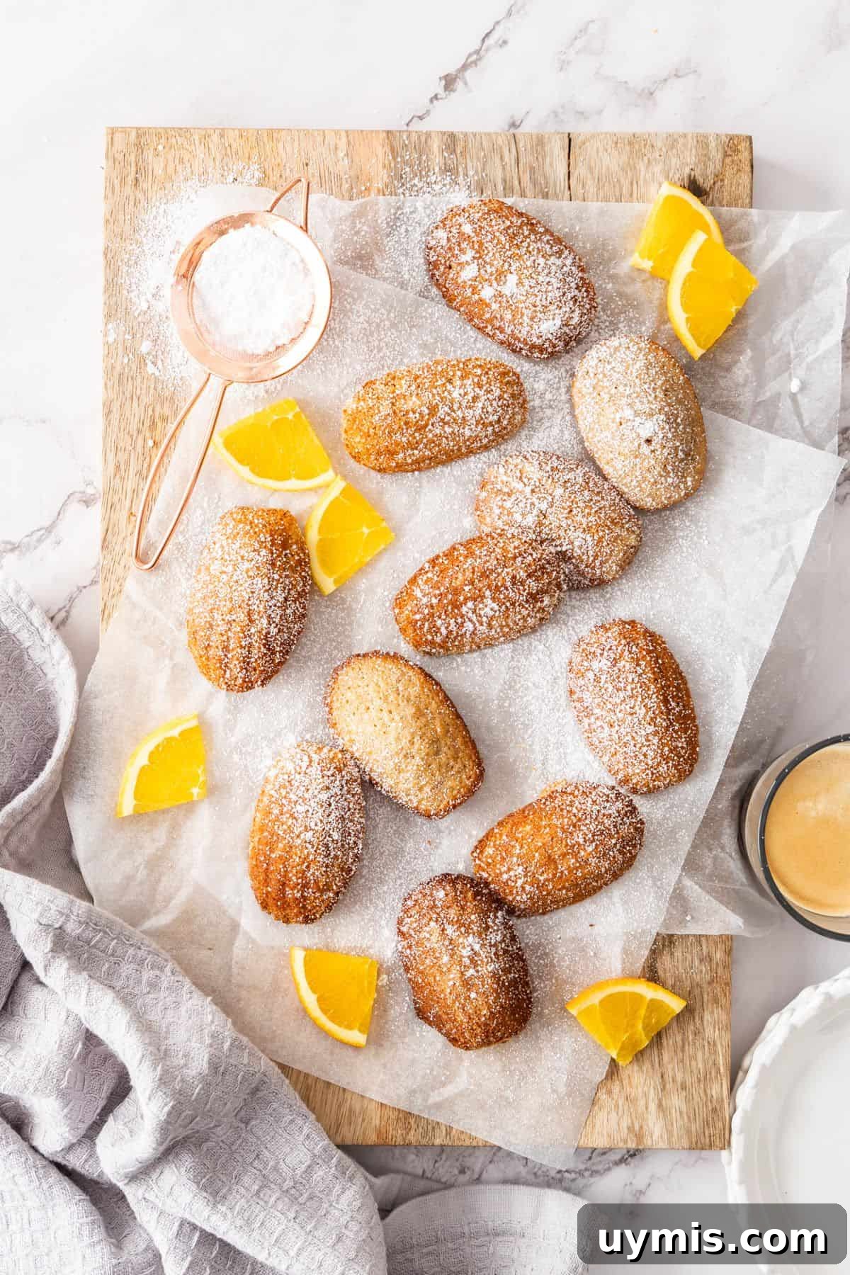 Spiced Orange Madeleines Recipe 4 Spiced Orange Madeleines on some baking paper on a wooden board, dusted with icing sugar and a few pieces of fresh orange.