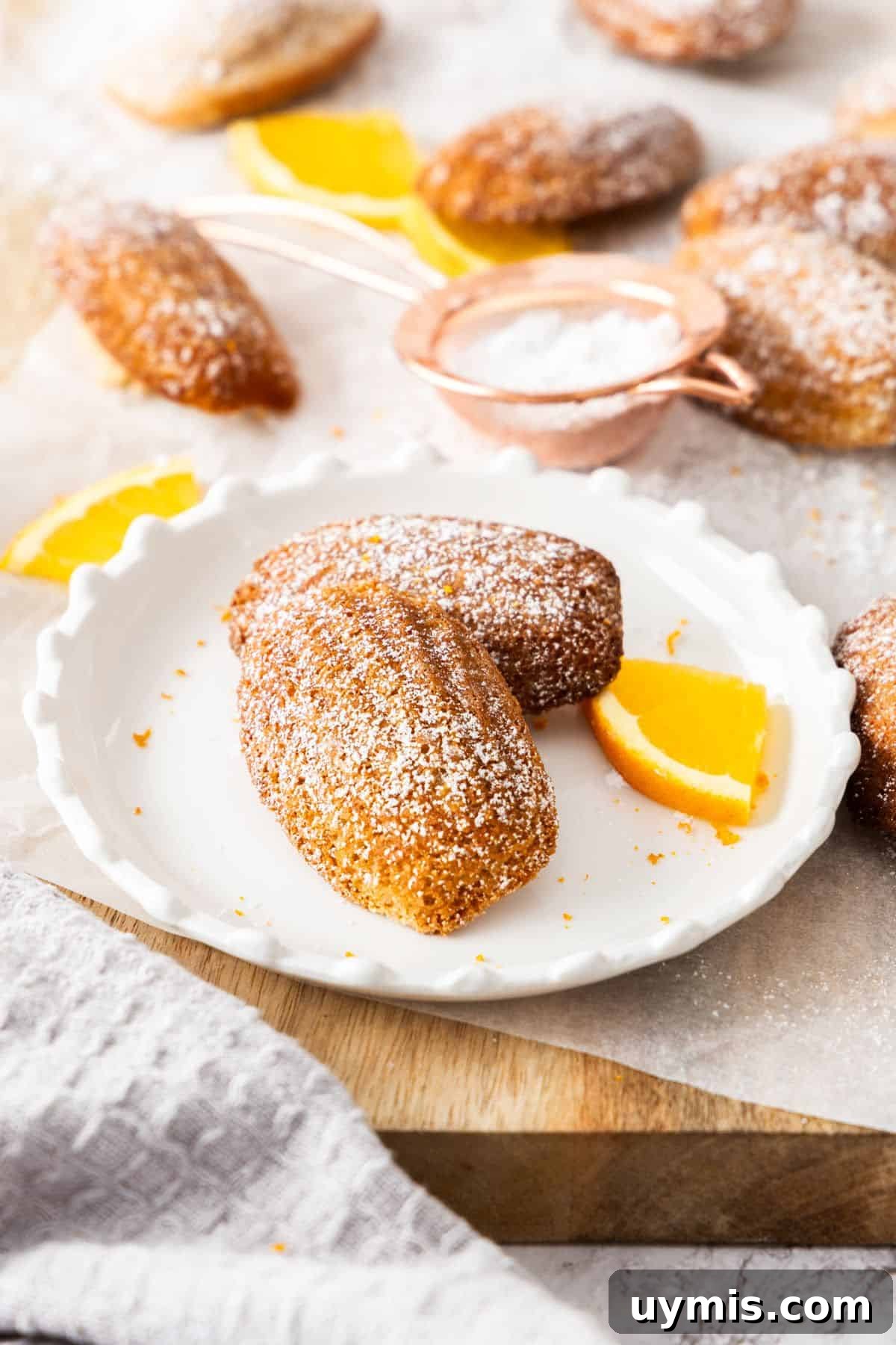 Spiced Orange Madeleines Recipe 8 Two Spiced Orange Madeleines sitting on a round white plate, on a board with other Madeleines around the edge.