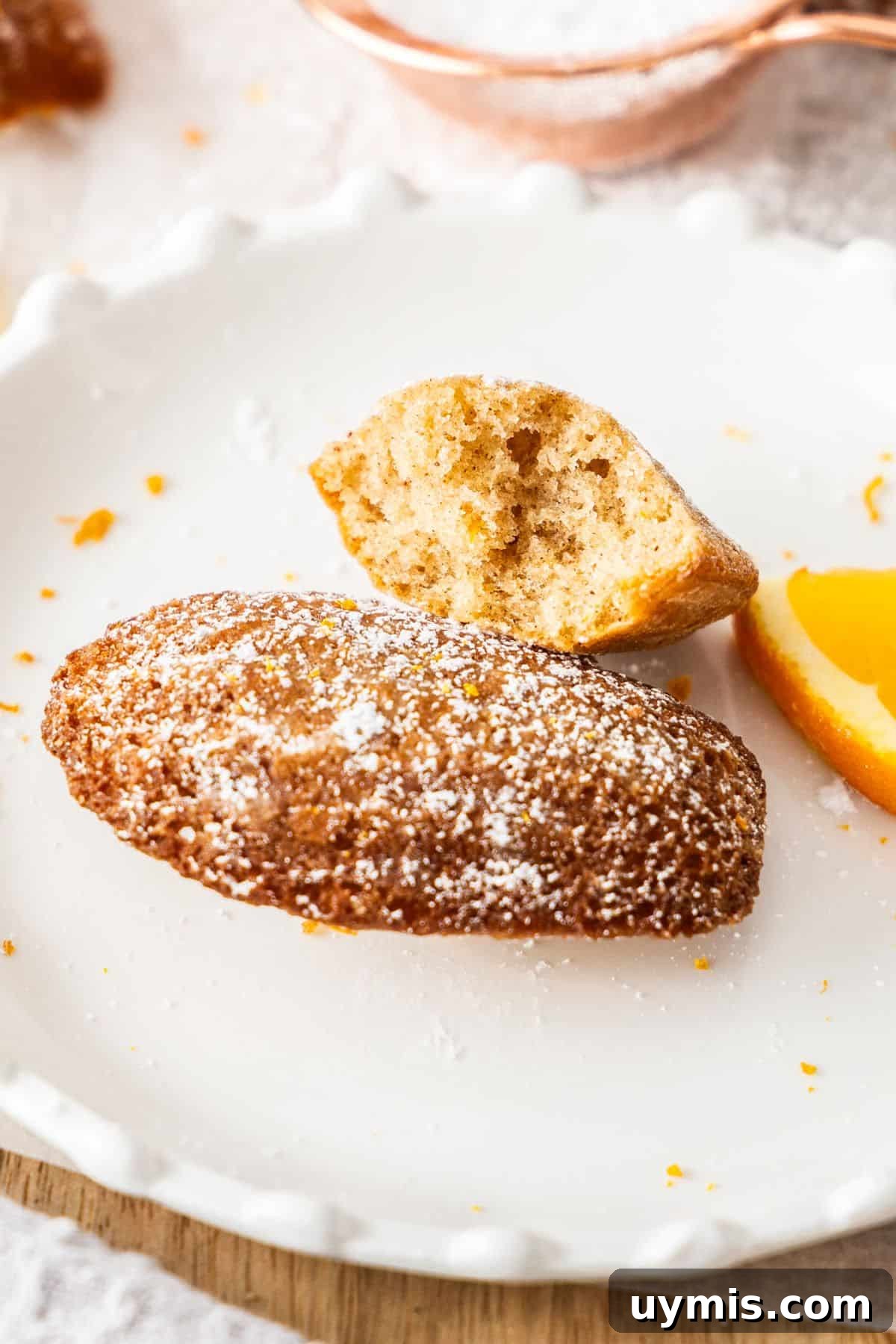 Spiced Orange Madeleines Recipe 9 Two Spiced Orange Madeleines sitting on a round white plate, one with a bite taken out of it to show the texture.