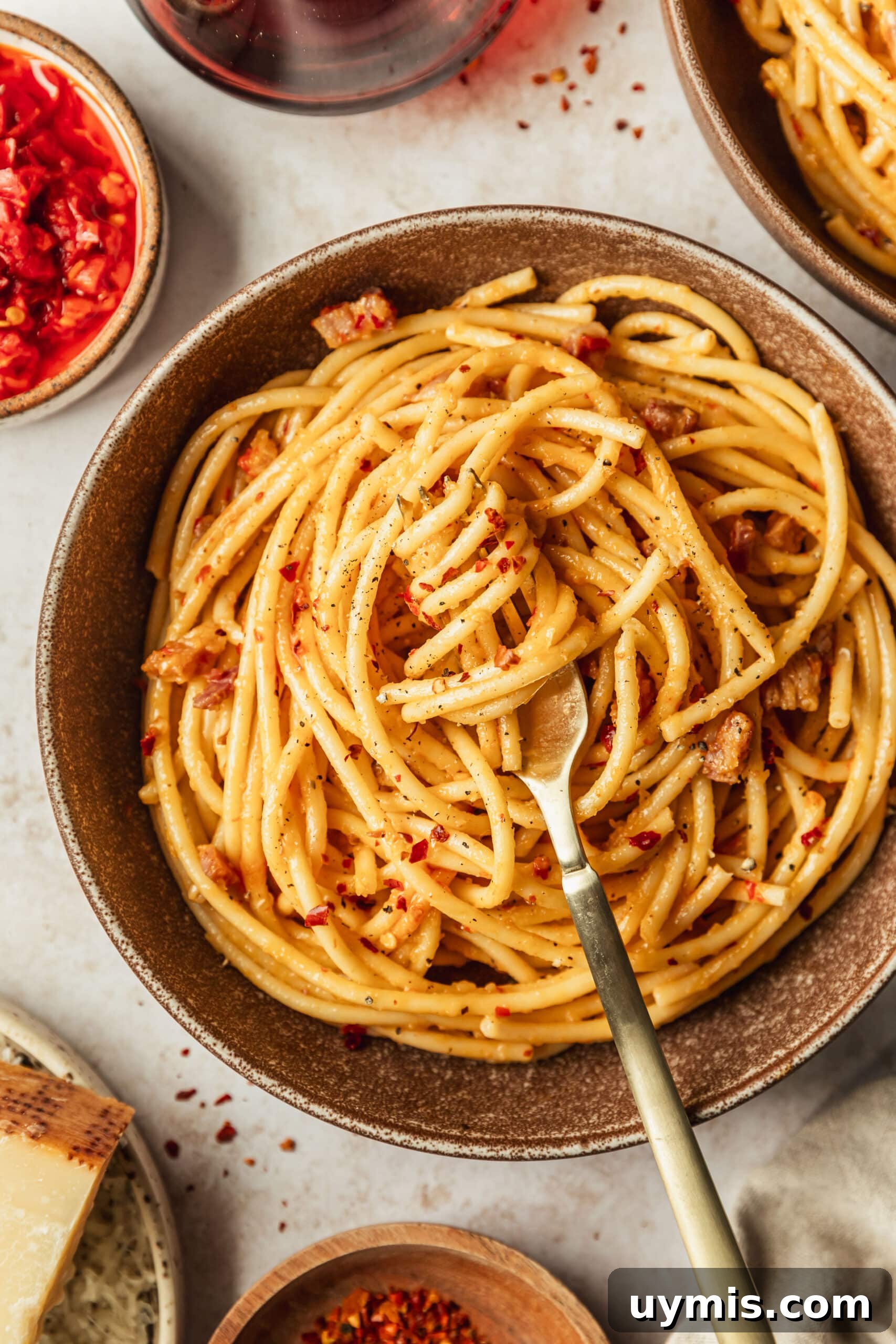 Creamy Spicy Bucatini Carbonara Recipe 2 A brown bowl of spicy bucatini carbonara on a tan counter next to glasses of red wine and brown bowls of parmesan, red pepper flakes, and Calabrian chili.