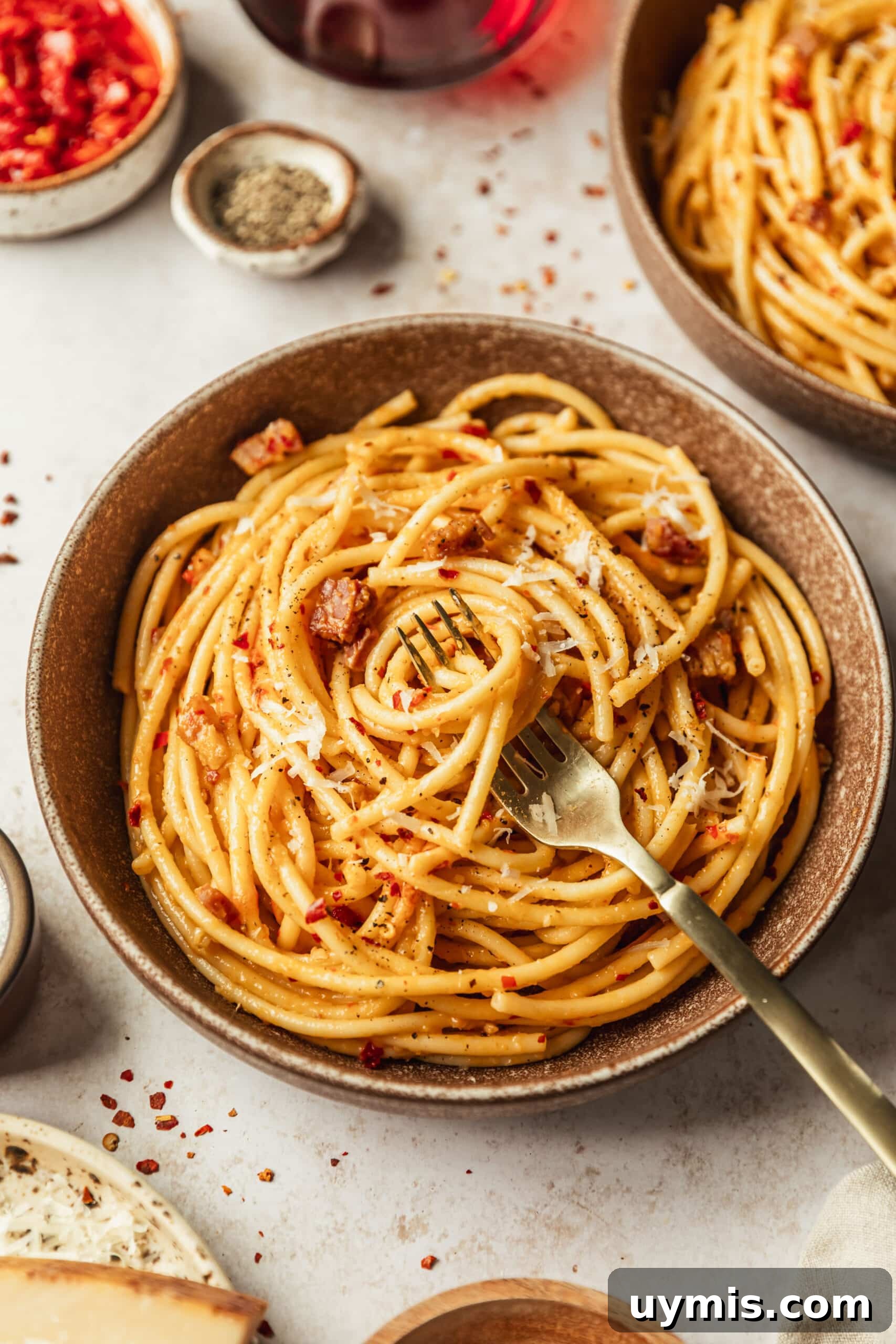 Creamy Spicy Bucatini Carbonara Recipe 11 Brown bowls of spicy bucatini alla carbonara on a tan counter next to white and brown bowls of chilis, pepper, and parmesan.