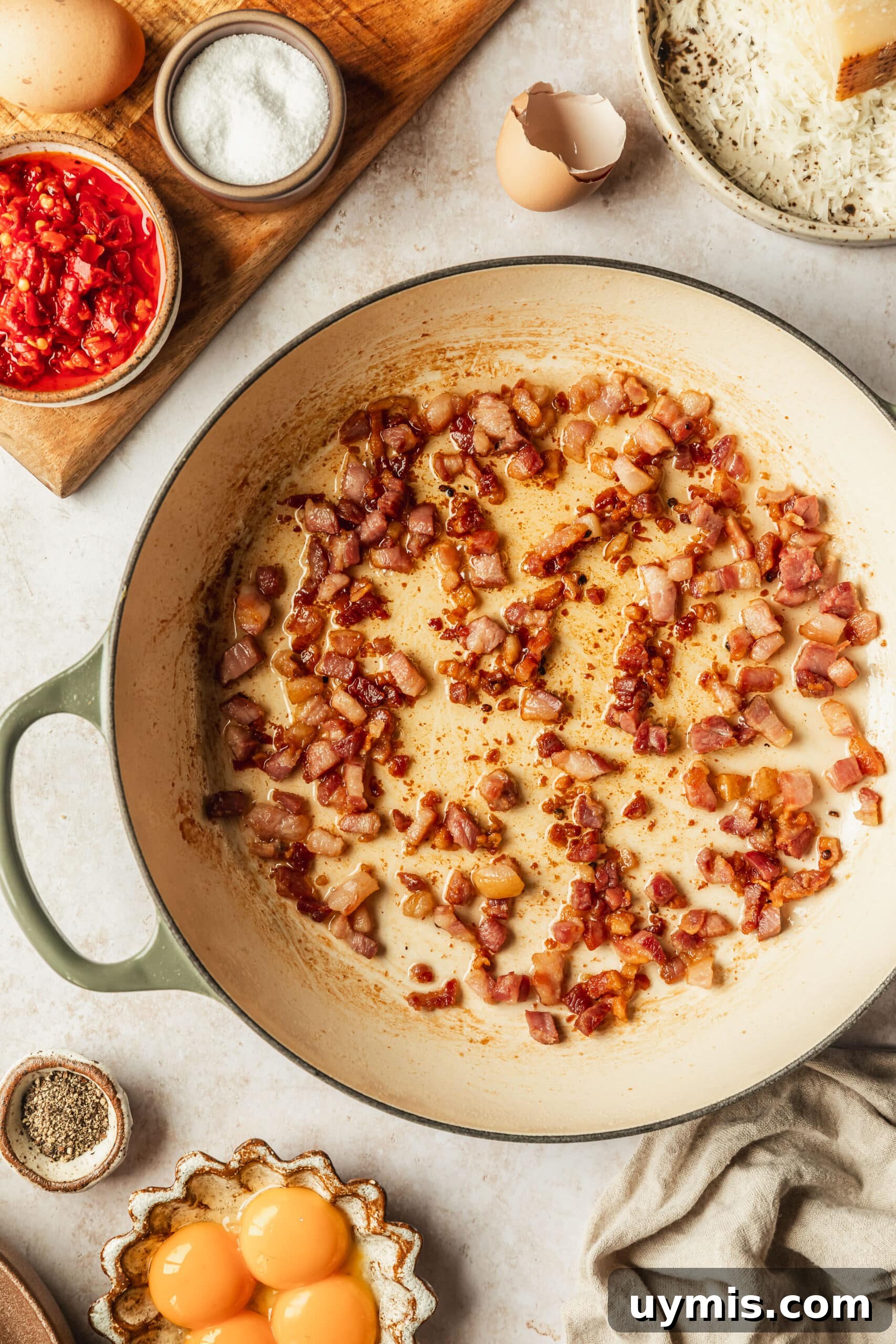 Creamy Spicy Bucatini Carbonara Recipe 6 A white pot of pancetta on a tan counter next to white and brown bowls of parmesan, chilis, eggs, salt, and pepper.