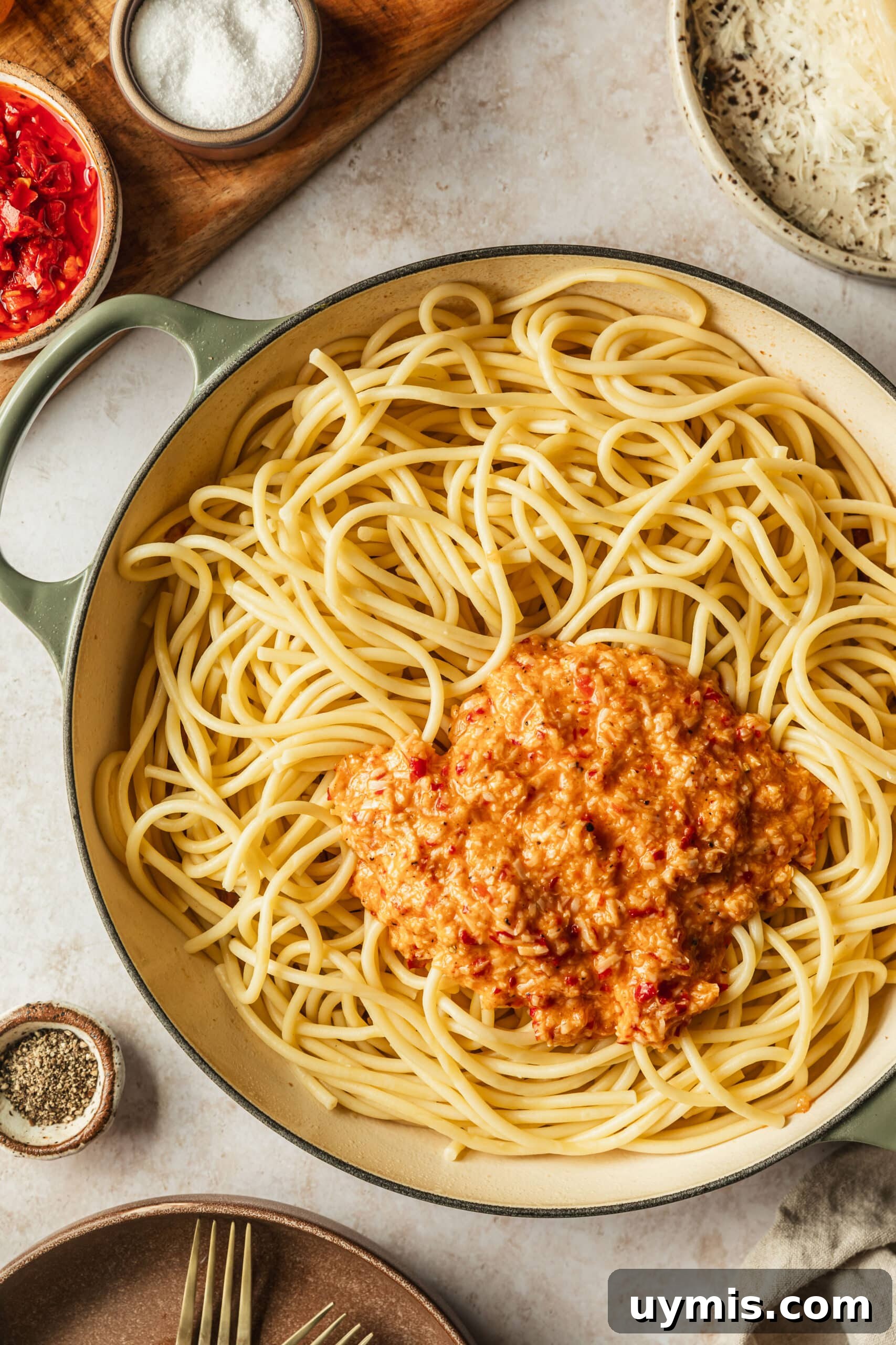 Creamy Spicy Bucatini Carbonara Recipe 9 A white pot of bucatini with sauce is on a tan counter next to brown bowls of salt, pepper, cheese, and chilis.
