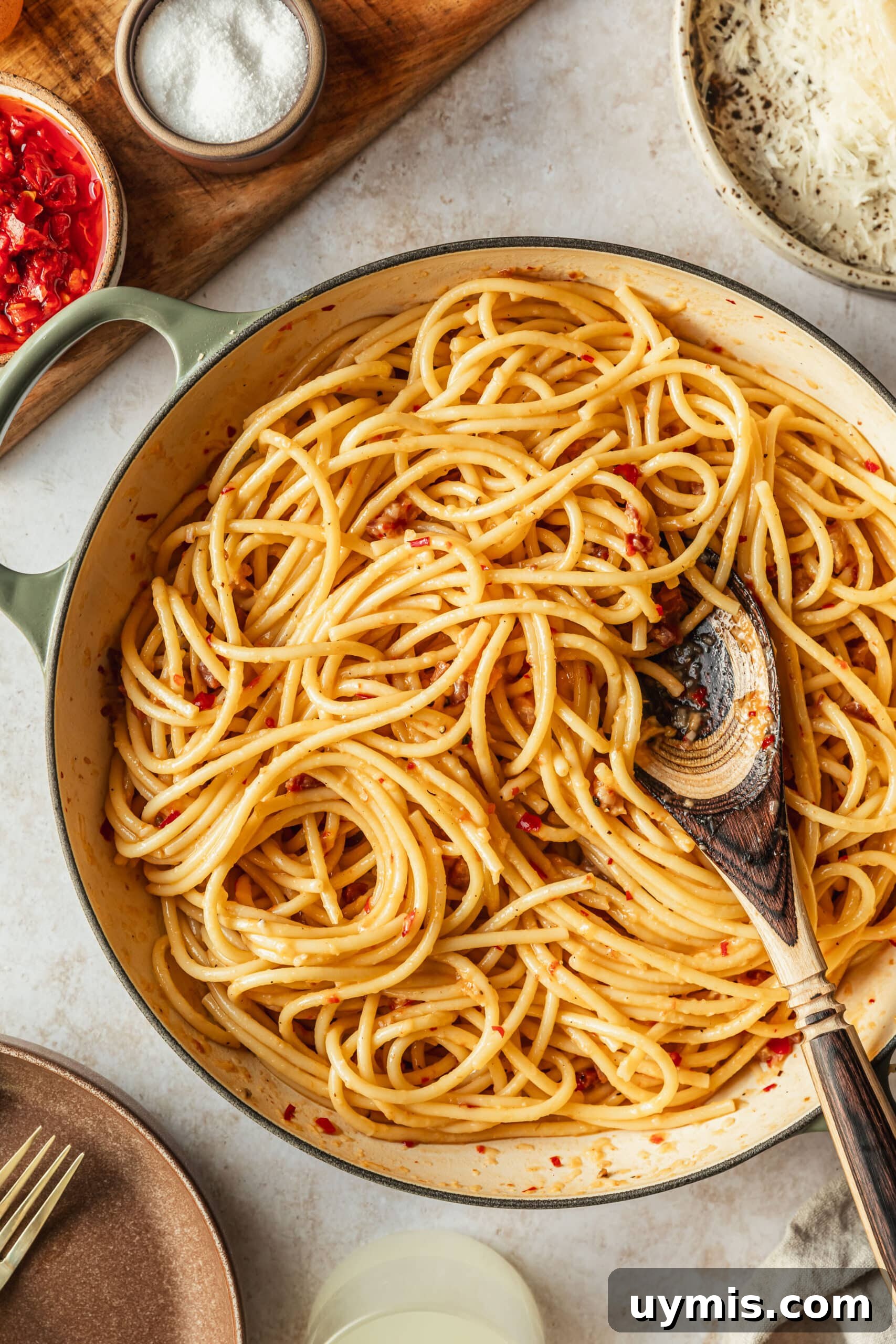 Creamy Spicy Bucatini Carbonara Recipe 10 A white pot of spicy bucatini carbonara on a tan counter next to brown bowls of cheese, chilis, salt, and pepper.