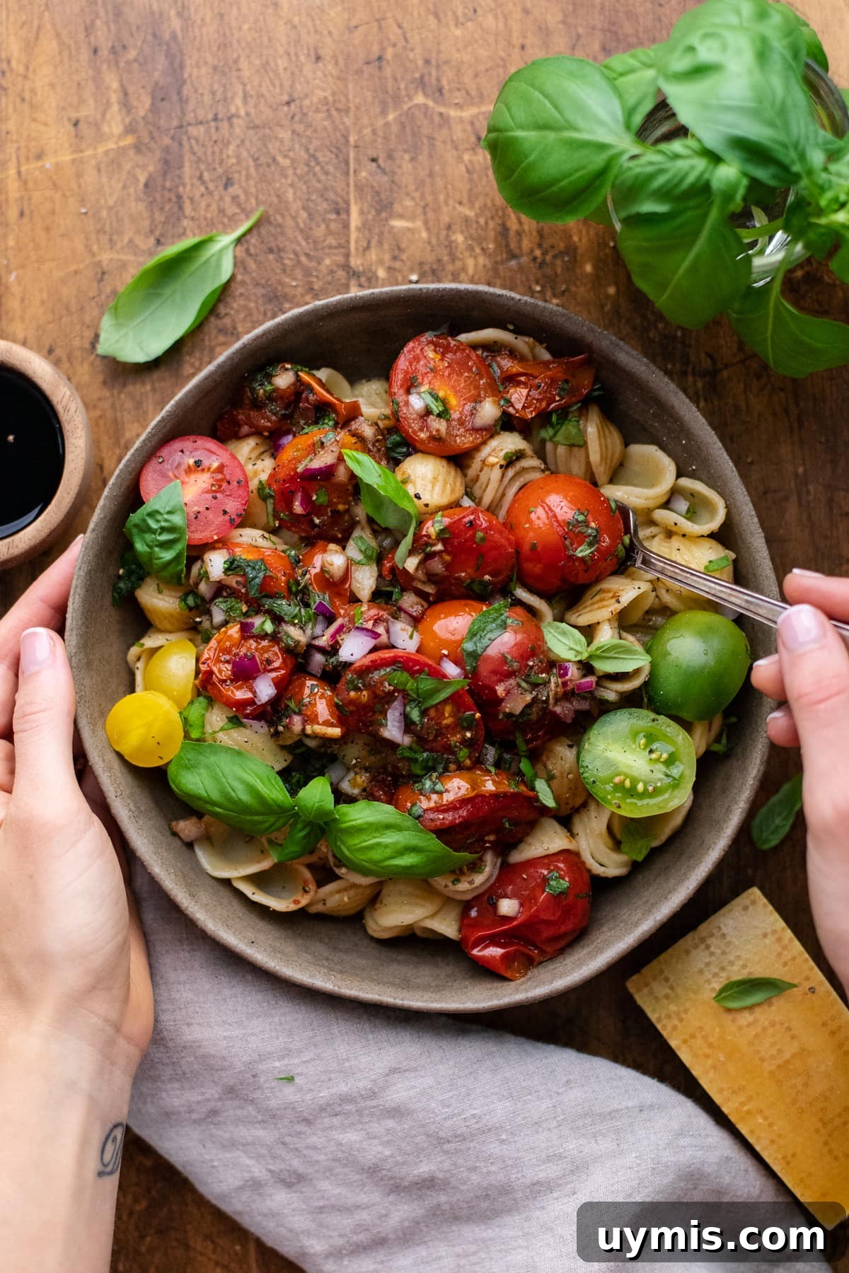 Bruschetta Pasta with Roasted Tomatoes and Basil 9 hands holding bowl with a fork in it of pasta with bruschetta in it on brown wooden board with herbs and gray napkin arranged around it.