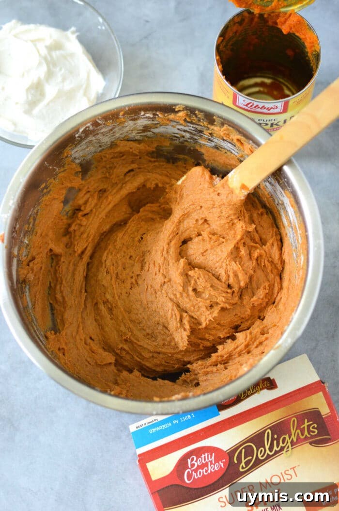 Moist Pumpkin Bars Recipe Made with Cake Mix 4 A mixing bowl filled with pumpkin cake bars batter sits on a gray surface, surrounded by canned pumpkin and a cake mix box, ready to whip up delicious pumpkin bars with cake mix. Nearby, whipped topping waits to add the finishing touch.