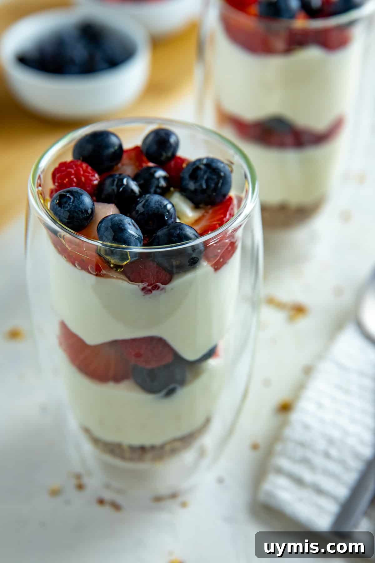 Layered Mixed Berry Parfaits with Greek Yogurt and Granola 2 Two tall clear glasses with a granola crust at the bottom, two layers of yogurt and two layers of strawberries and blueberries.