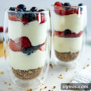 Two tall clear glasses with a granola crust at the bottom, two layers of yogurt and two layers of strawberries and blueberries.