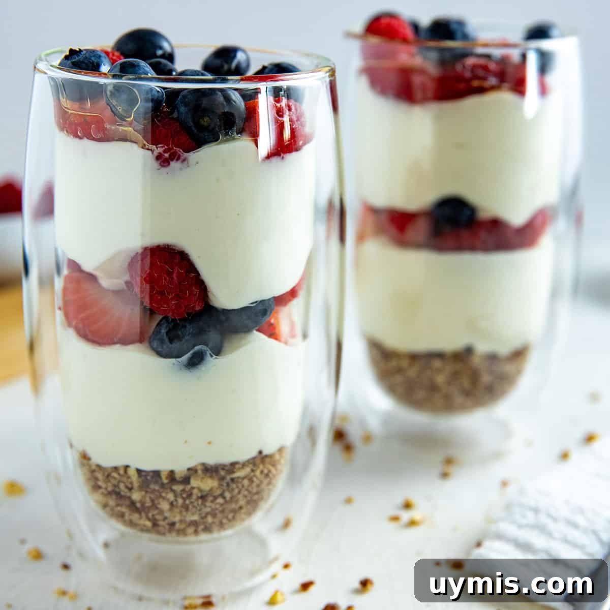 Layered Mixed Berry Parfaits with Greek Yogurt and Granola 7 Two tall clear glasses with a granola crust at the bottom, two layers of yogurt and two layers of strawberries and blueberries.