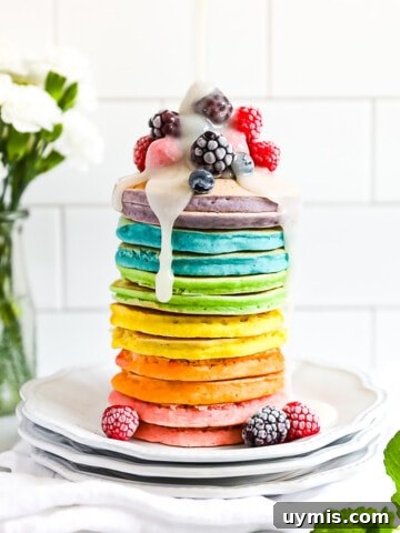 A stack of colorful pancakes with frozen fruit on top and a drizzle of white sugar sauce on top.