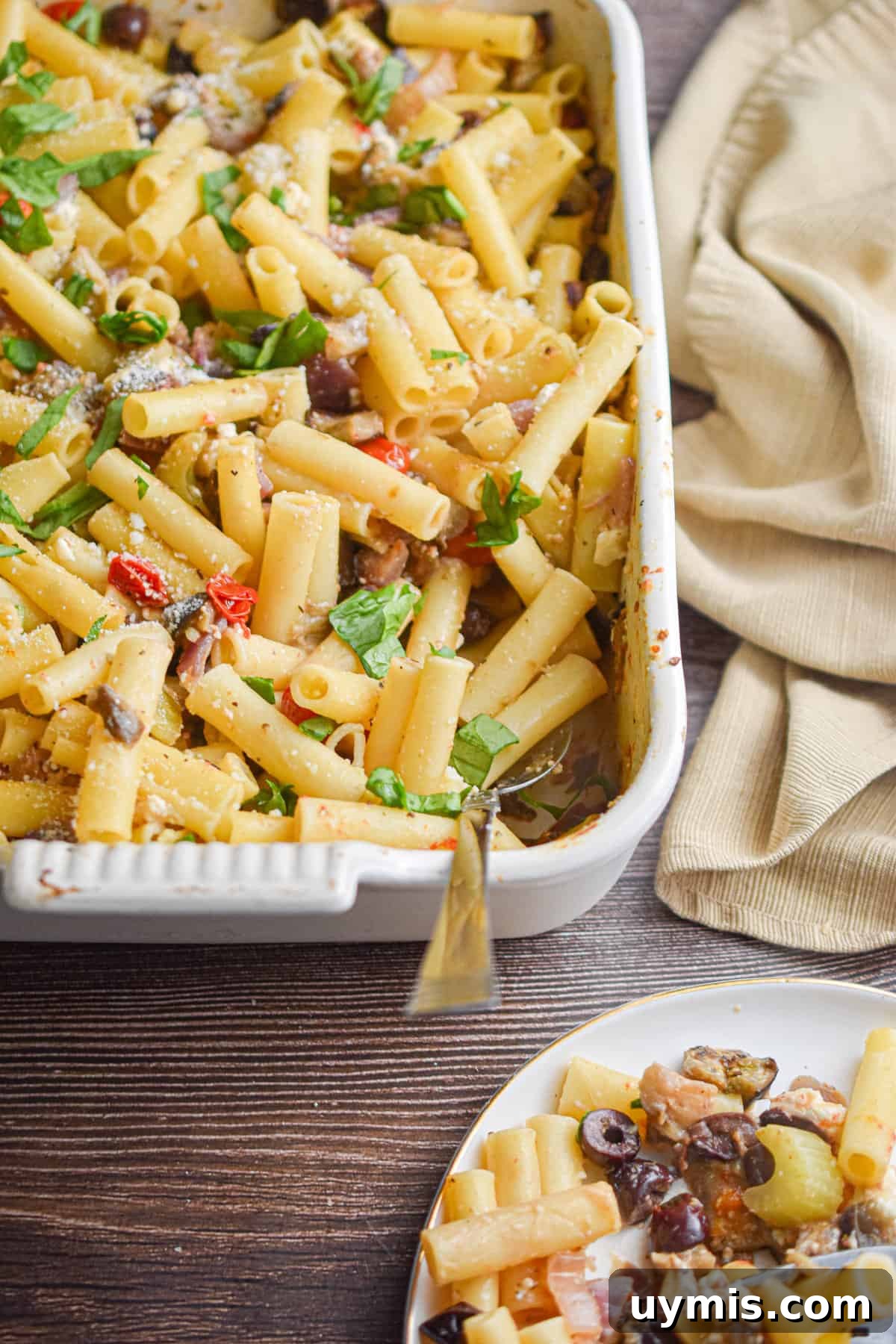 Creamy Greek Baked Pasta with Feta 2 A napkin is behind the baking dish with the Greek Pasta Bake in it and a portion is on a plate in front of the baking dish.