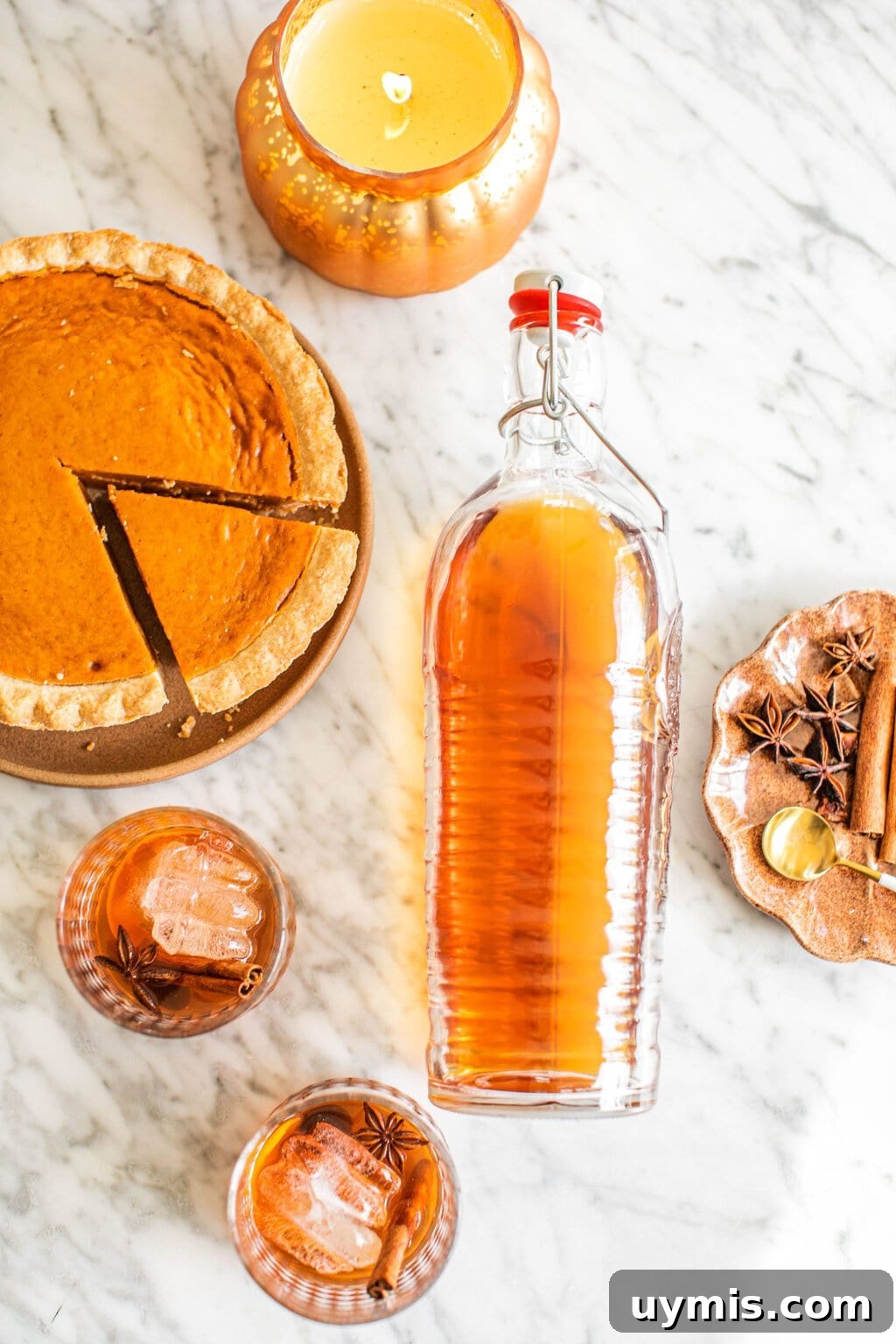 an overhead shot of two glasses of old fashioneds, a large bottle of the cocktail, a pumpkin pie, and garnish