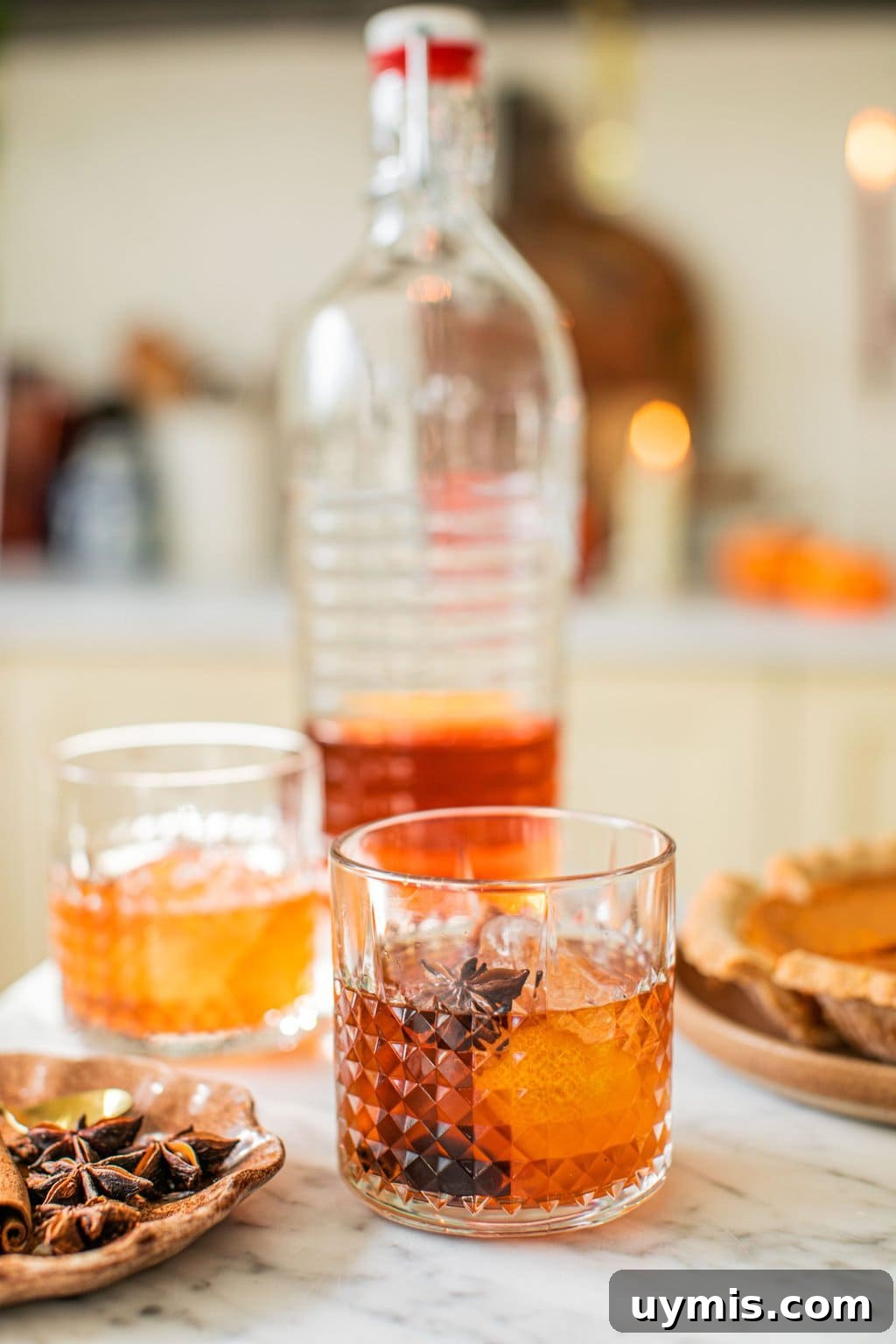 two glasses of old fashioneds beside a plate with garnish on it and a large bottle full of the cocktail and a pumpkin pie out of focus in the background