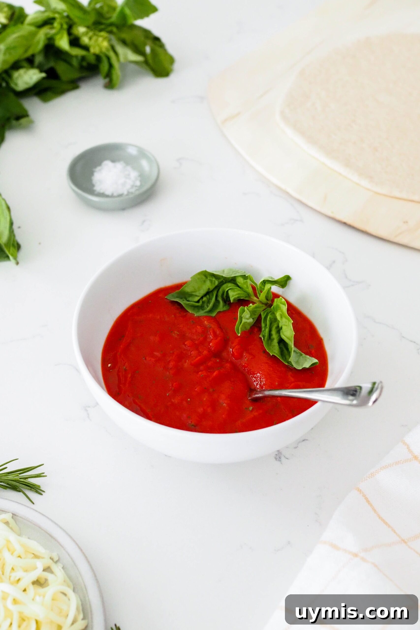 How to Make Pizza Sauce from Tomato Paste 2 A bowl of pizza sauce next to a bunch of basil and a rolled out pizza crust.