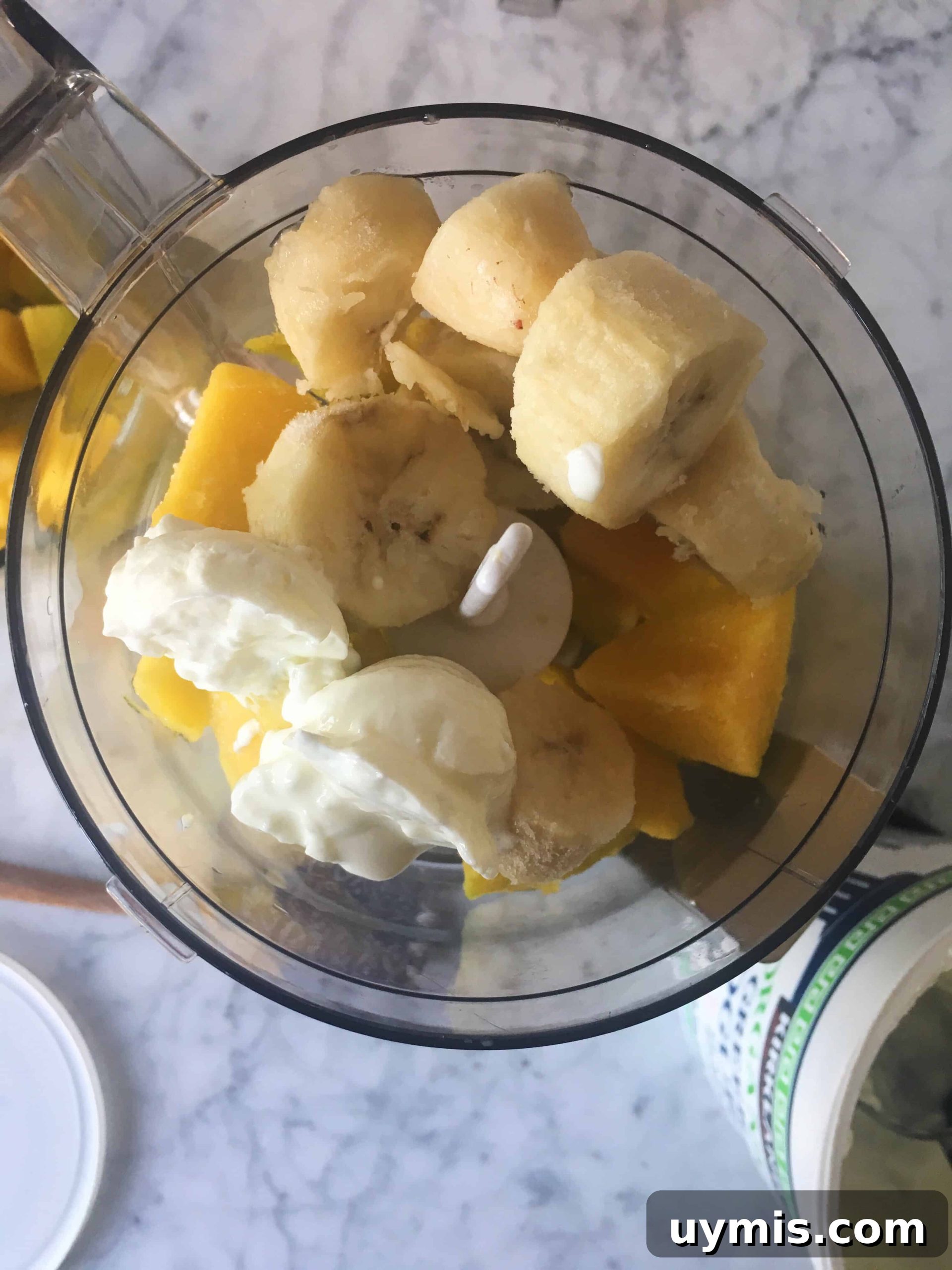 Bananas and mangoes with greek yogurt in a food processor about to be blended to make the smoothie bowl