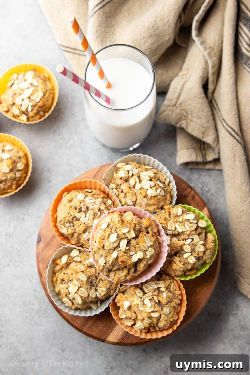 Wholesome Banana Oat Muffin Recipe 4 Healthy banana oat muffins in colorful muffin liners on a wooden stand with a white and black tea towel and a glass of milk with two straws.