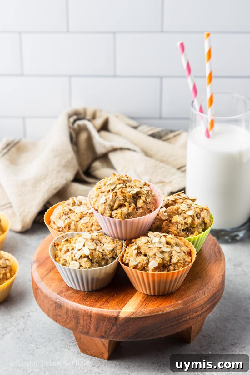 Wholesome Banana Oat Muffin Recipe 5 Healthy banana oat muffins in colorful muffin liners on a wooden stand with a white and black tea towel and a glass or milk with two straws.