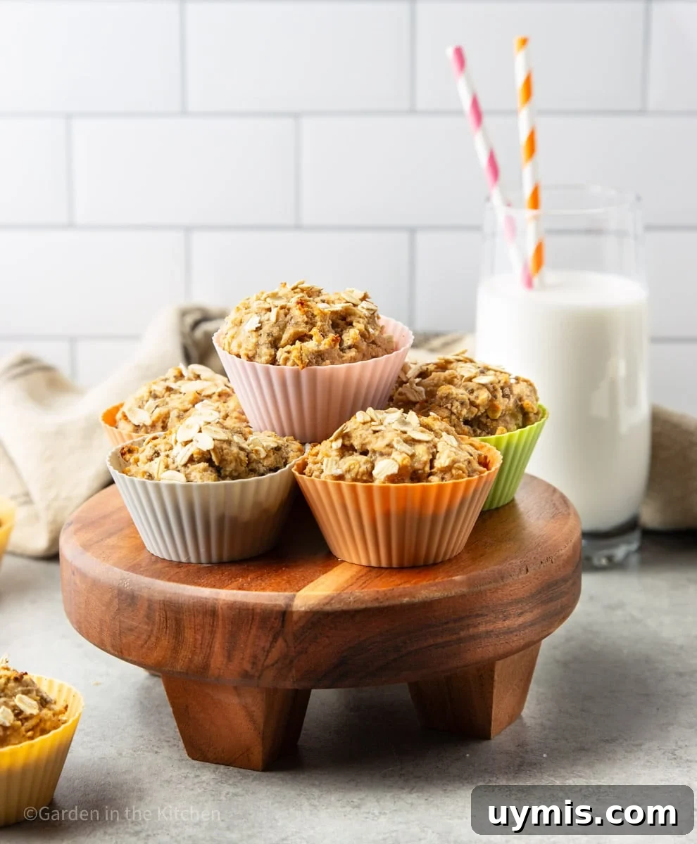 Wholesome Banana Oat Muffin Recipe 6 Healthy banana oat muffins in colorful muffin liners on a wooden stand with a white and black tea towel and a glass or milk with two straws.