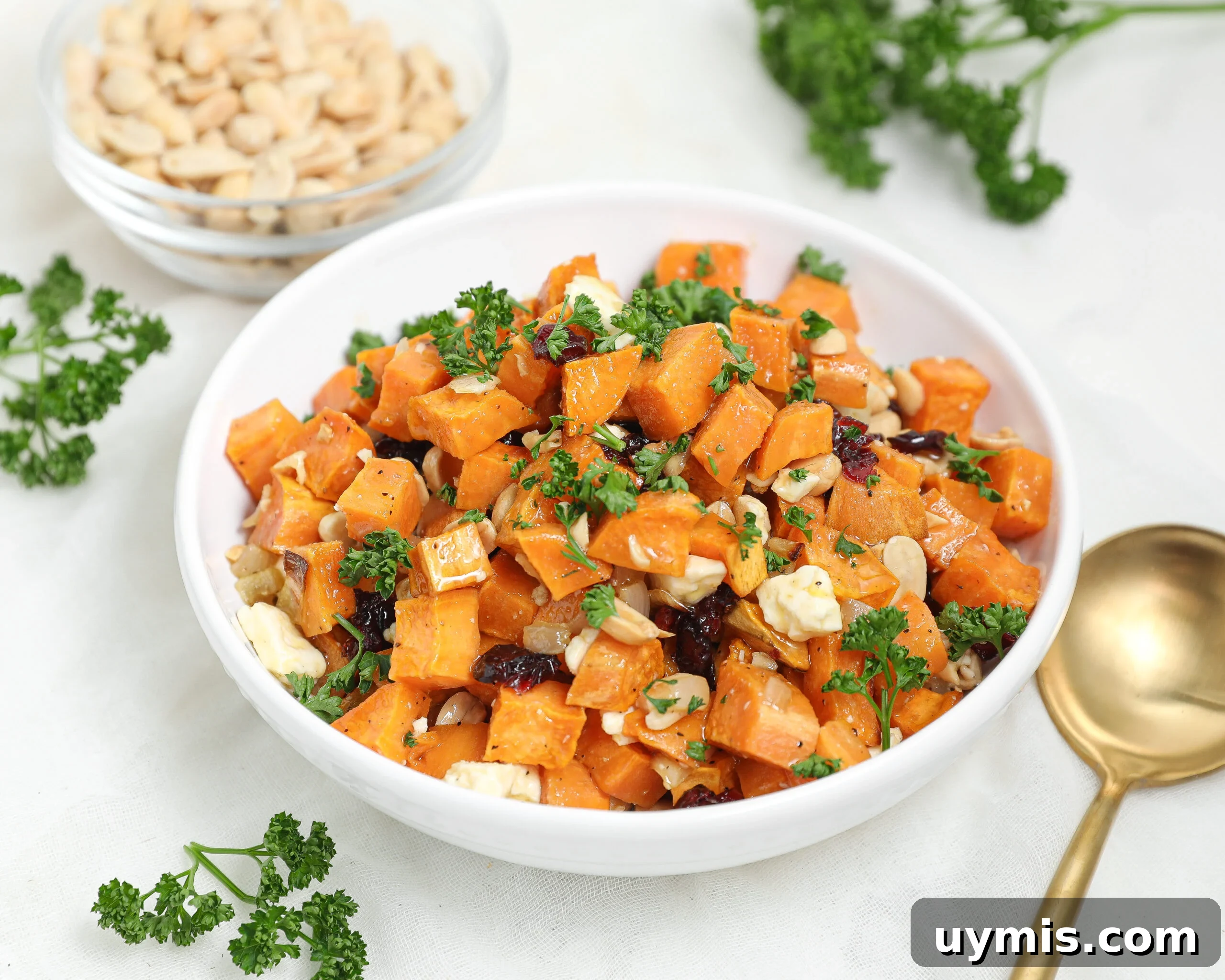 Maple Roasted Sweet Potatoes with Bacon and Walnuts 11 A white bowl filled with a salad of cubed roasted sweet potatoes, chopped parsley, nuts, and crumbled cheese, with a gold spoon, fresh parsley, and a bowl of nuts nearby on a light surface.