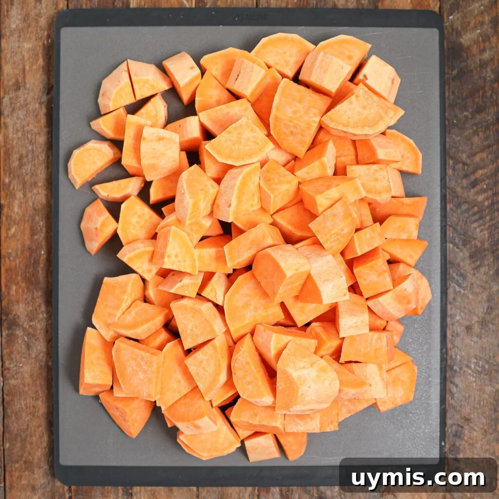 Maple Roasted Sweet Potatoes with Bacon and Walnuts 3 Cubed sweet potatoes are spread out on a dark cutting board, ready to become Maple Roasted Sweet Potato with Bacon & Walnuts. The orange cubes sit neatly on a rustic wooden surface, perfect for cooking or roasting.