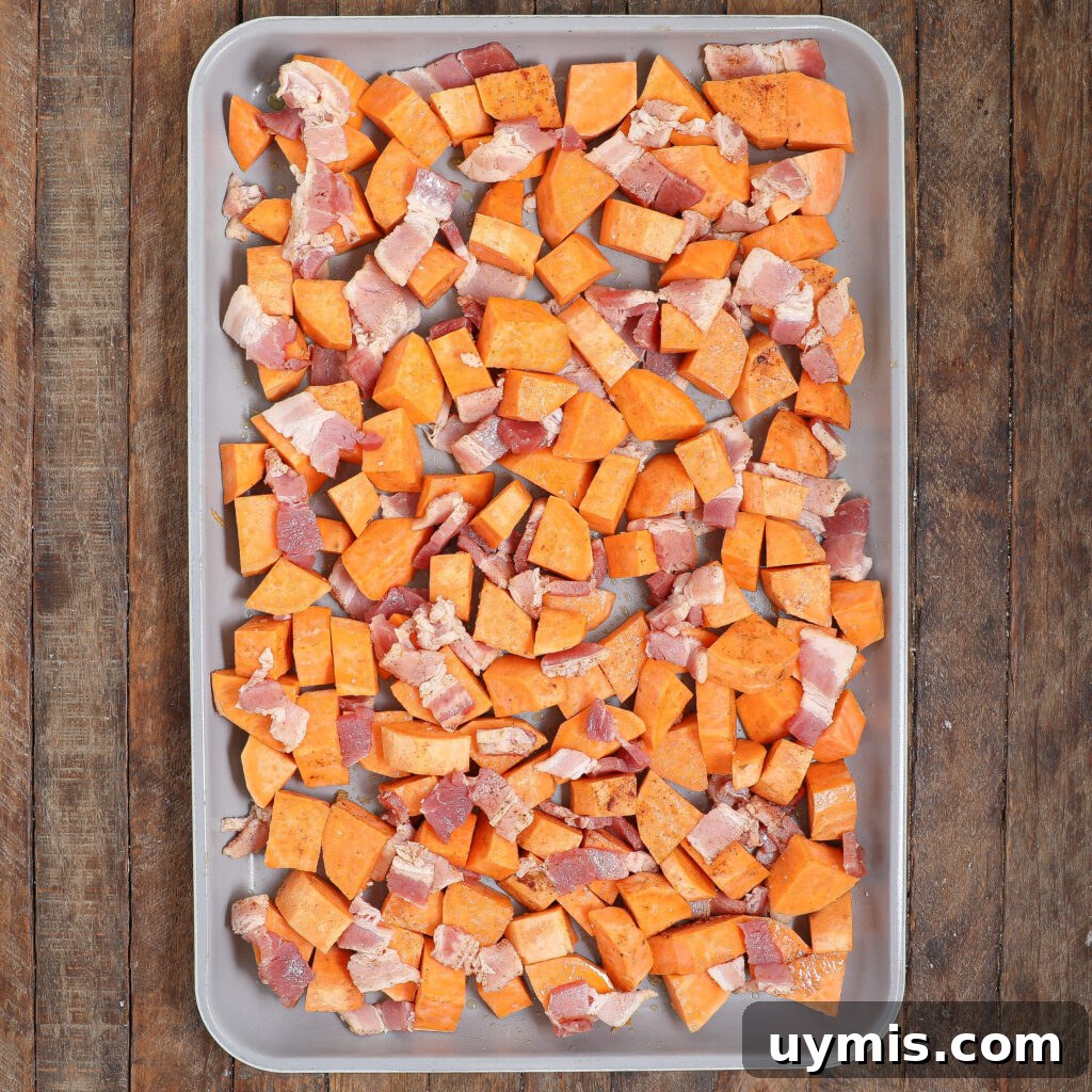 Maple Roasted Sweet Potatoes with Bacon and Walnuts 5 A baking sheet filled with uncooked diced sweet potatoes and chunks of raw bacon, arranged for a Maple Roasted Sweet Potato with Bacon & Walnuts dish, sits in an even layer on a wooden surface.