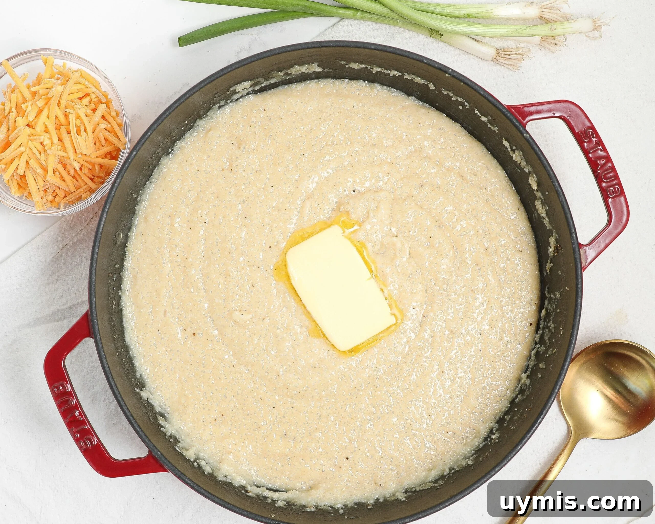 Maple Roasted Sweet Potatoes with Bacon and Walnuts 9 A red pot filled with creamy grits topped with a pat of melting butter. Beside the pot are a bunch of green onions, a gold ladle, and a small bowl of shredded cheddar cheese on a white surface.