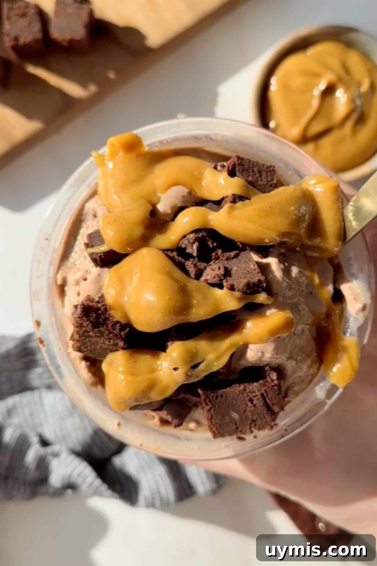 Chocolate Brownie Protein Ice Cream Recipe for the Creami 2 A cup of chocolate ice cream topped with caramel sauce and brownie chunks, with a spoon in it. A bowl with caramel is visible in the background.