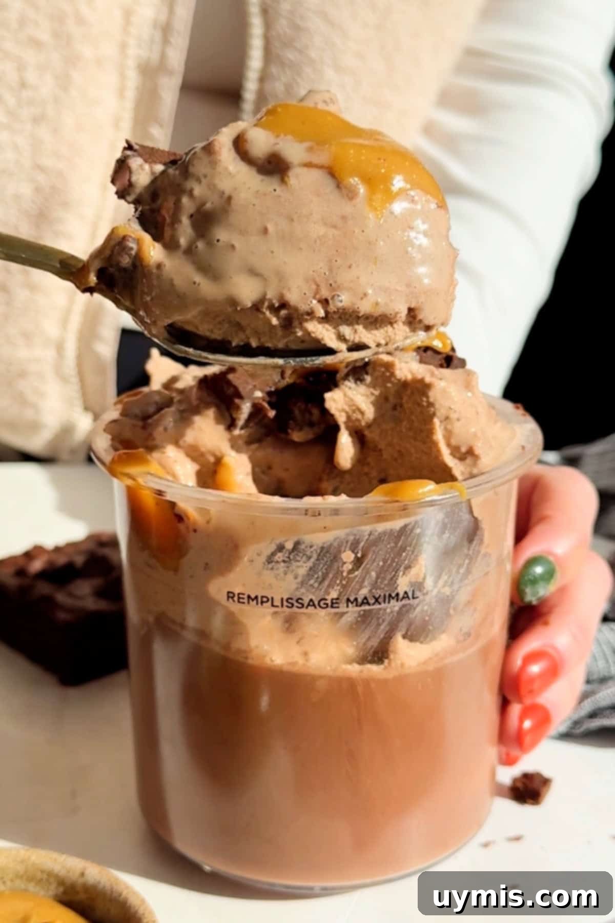 Chocolate Brownie Protein Ice Cream Recipe for the Creami 4 A person holds a spoon with ice cream drizzled with caramel over a clear cup, which seems filled to the maximum line. A brownie is visible in the background.