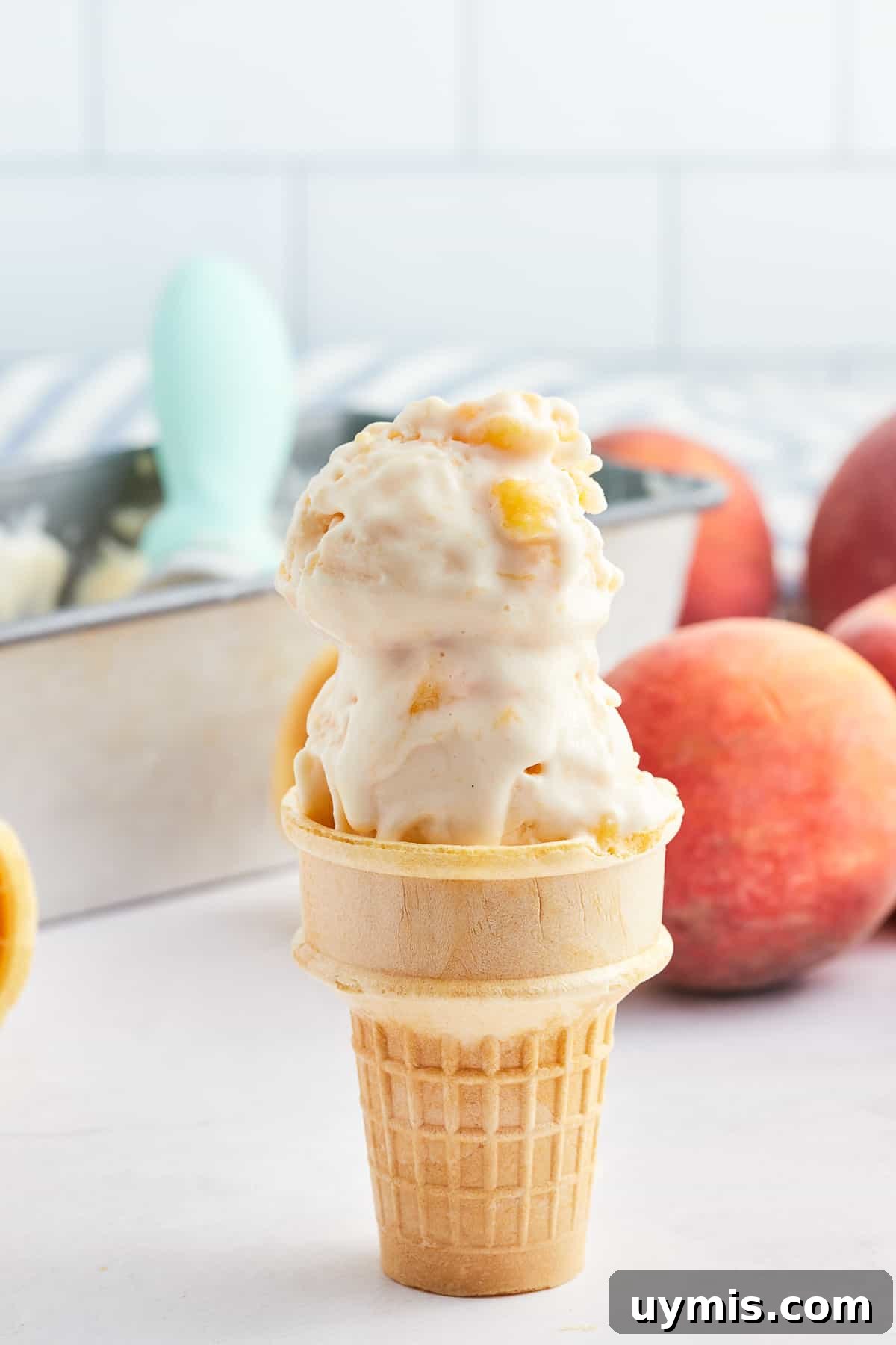 Homemade Peach Frozen Yogurt Recipe 7 Two scoops of peach frozen yogurt on an ice cream cone in front of fresh peaches.