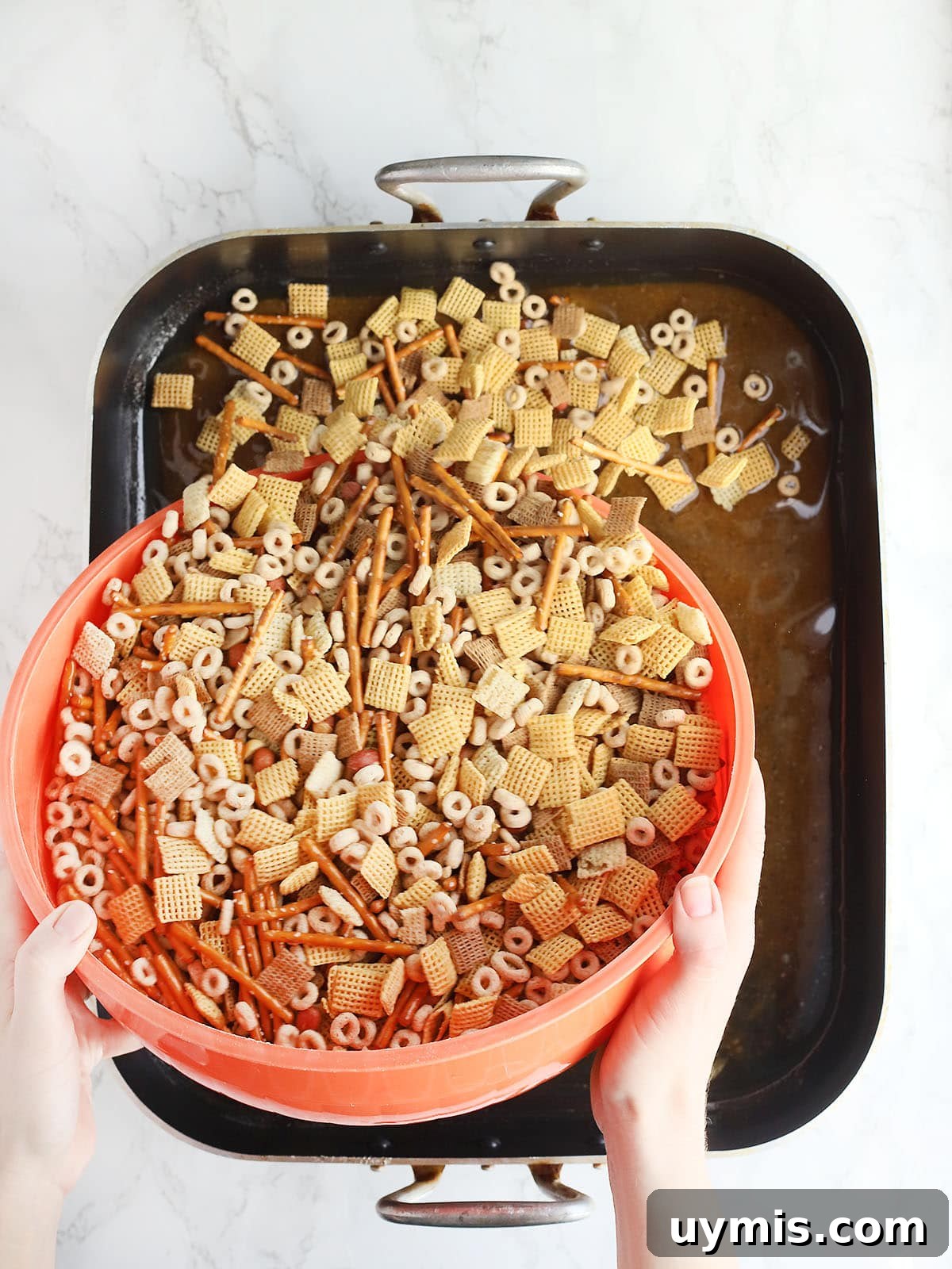 Crispy Oven Baked Chex Mix with Bacon Drippings 6 Hand pouring an orange bowl of mixed chex mix ingredients into a roasting pan of melted butter and Worcestershire sauce.