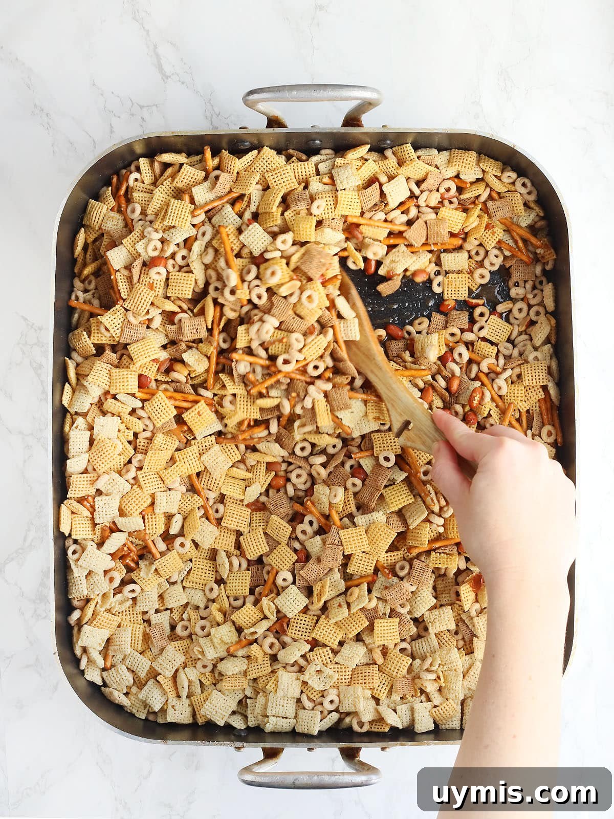 Crispy Oven Baked Chex Mix with Bacon Drippings 7 Hand using a wooden spoon to stir Chex mix in a large roasting pan.