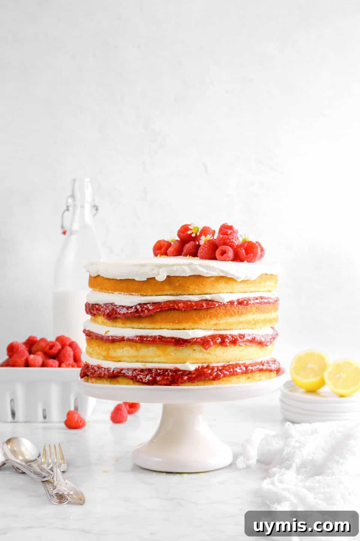 lemon raspberry jam cake on white cake plate with cheese cloth, forks, sliced lemon, raspberries, and milk