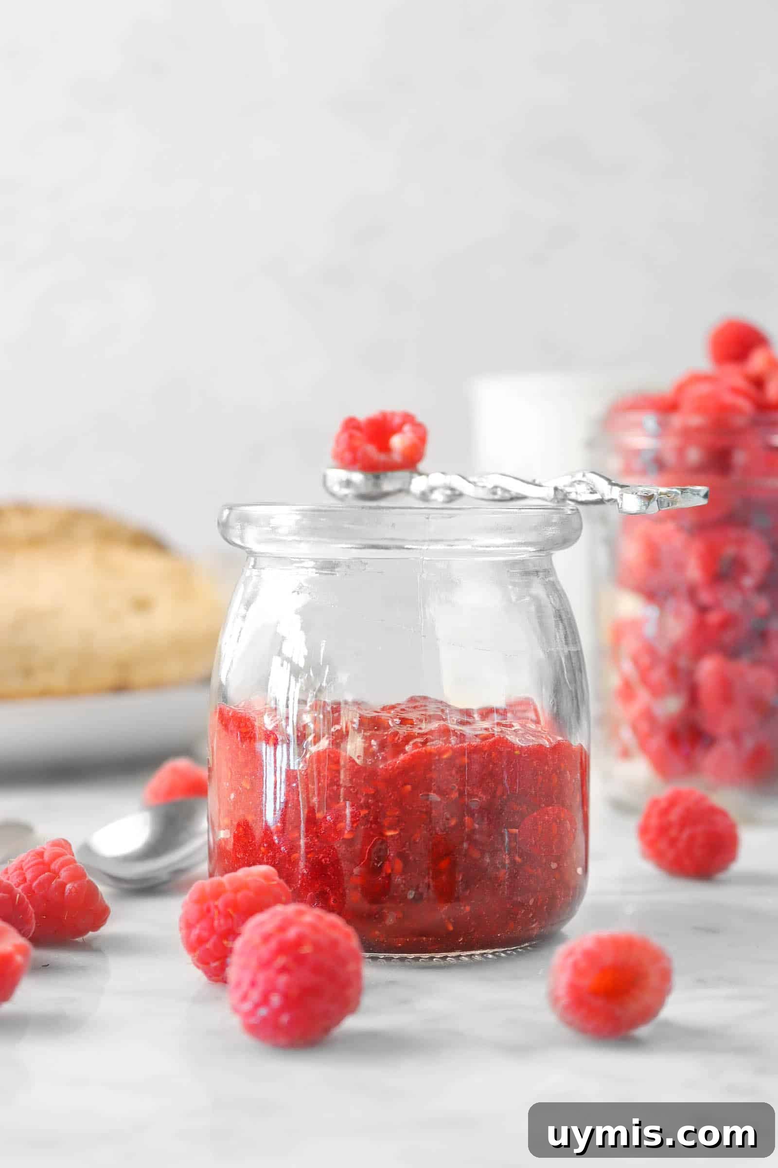 raspberry jam in a glass jar with fresh raspberries, bread, and a jar of raspberries in the background