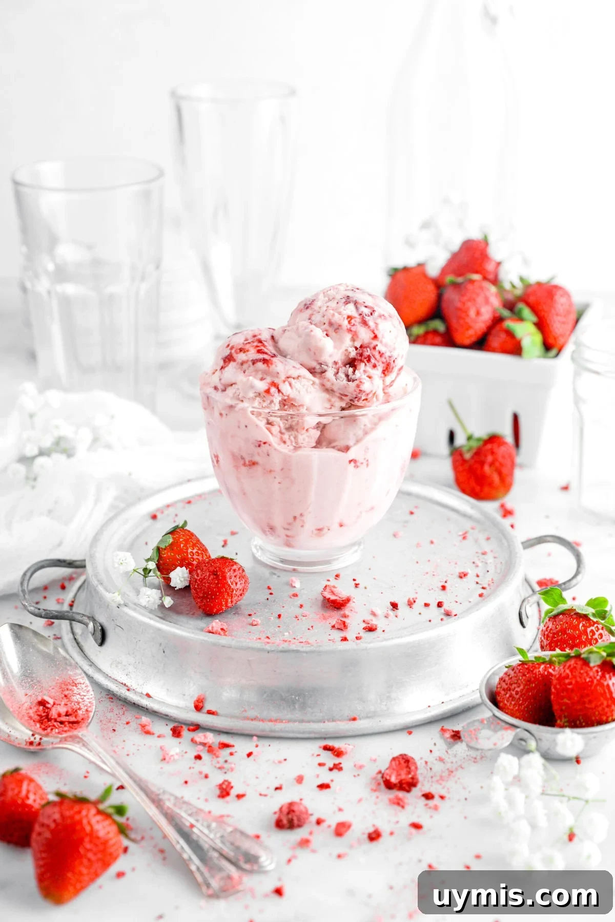 strawberry ice cream in glass cup on top of upside down cake pan with spoons, flowers, and strawberries around.