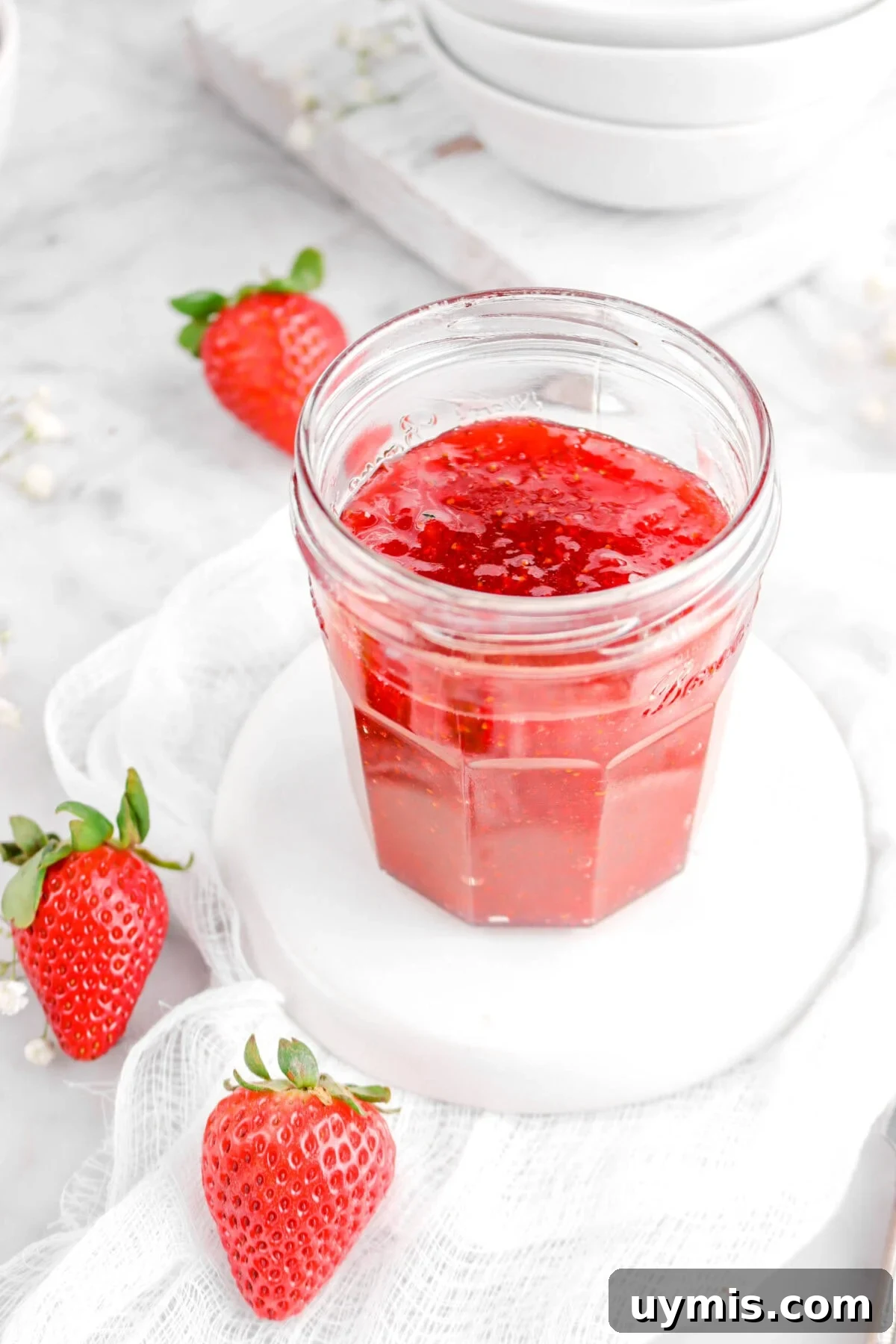 angled photo of strawberry jam on white upside down plate with three fresh strawberries beside and white cheese cloth